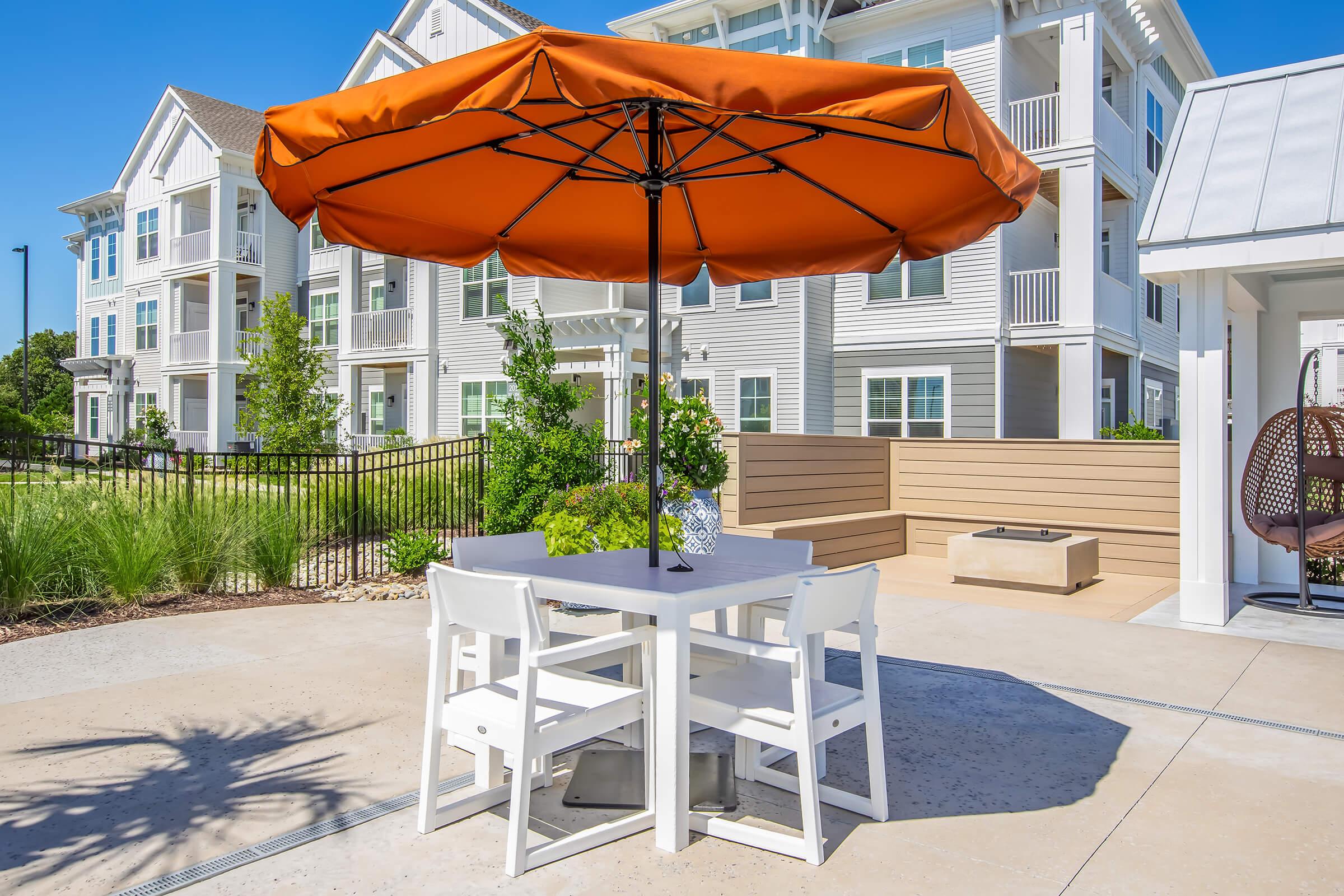 A patio area featuring a white table and chairs under a large orange umbrella. The background includes several modern apartment buildings and landscaped greenery, creating a bright and inviting outdoor space.