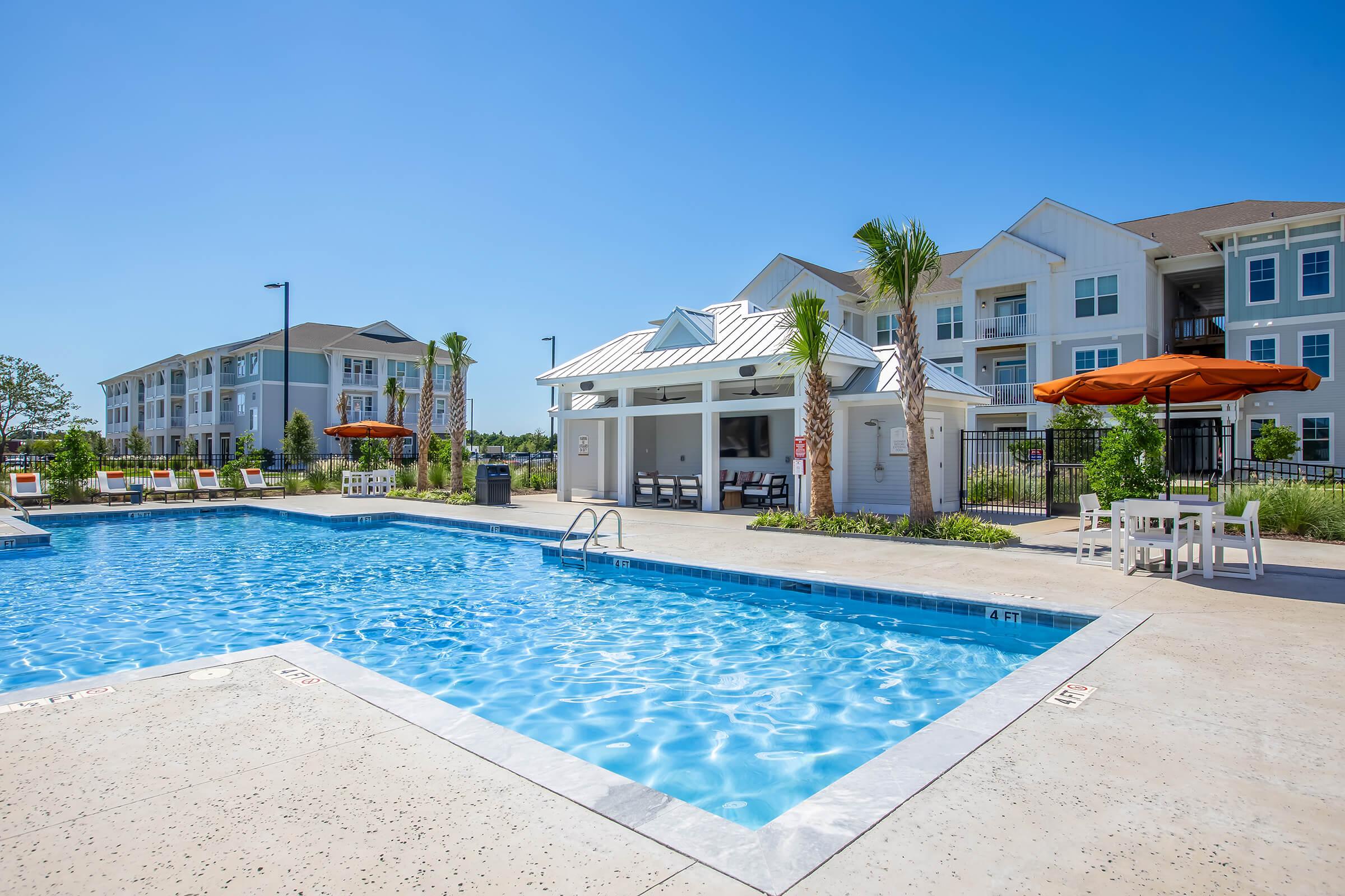A bright outdoor pool area featuring clear blue water, lounge chairs, and orange umbrellas. Surrounding the pool are palm trees and a well-maintained landscape, with an adjacent building displaying modern architecture in the background. The sky is clear and blue, emphasizing a beautiful day.