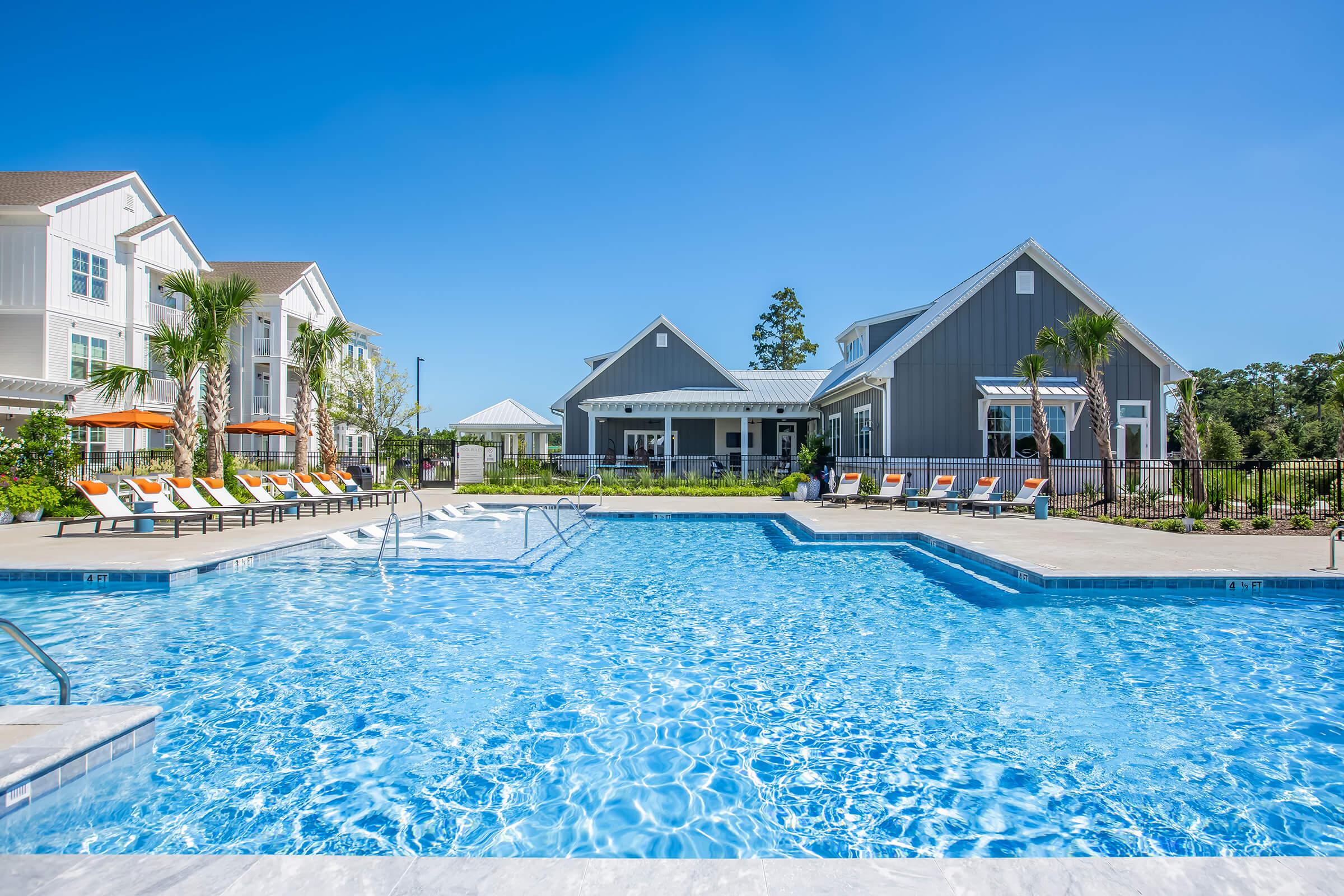 A sunny outdoor pool area featuring a large, clear blue pool surrounded by lounge chairs and palm trees. In the background, there's a modern building with a gray exterior, and sunny skies overhead. The scene conveys a relaxing atmosphere perfect for leisure and enjoyment.