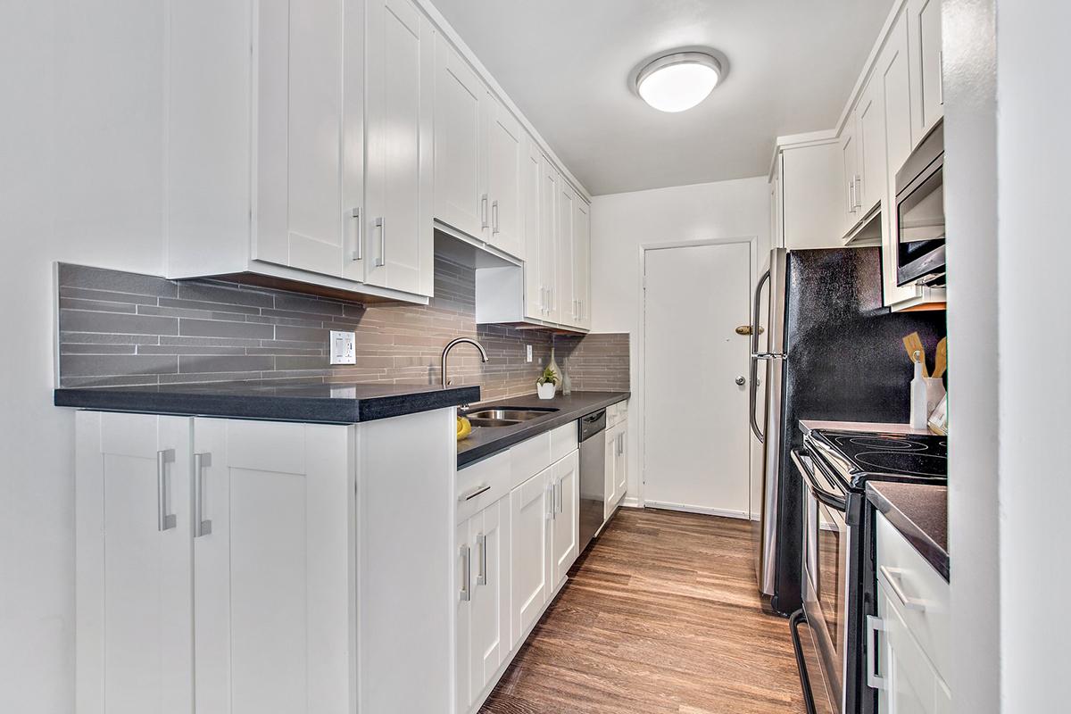 Modern kitchen featuring white cabinets, a dark countertop, stainless steel appliances, and a tiled backsplash. The layout includes a single sink and a doorway leading out of the kitchen, with wood-like flooring enhancing the contemporary design.