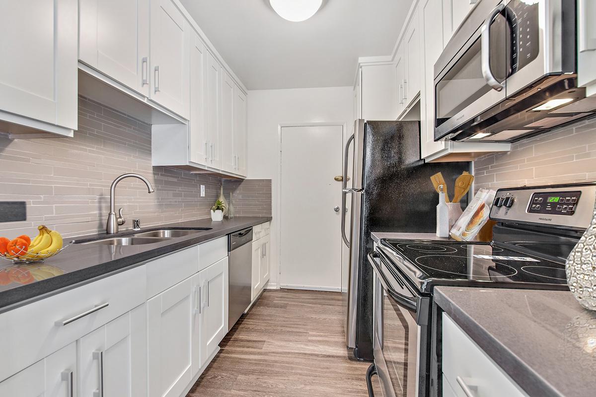 A modern kitchen featuring white cabinets, stainless steel appliances, and black countertops. The space includes a double sink, a stove, and a microwave, along with a bowl of bananas on the counter. Light gray backsplash tiles complement the design, creating a bright and inviting atmosphere.