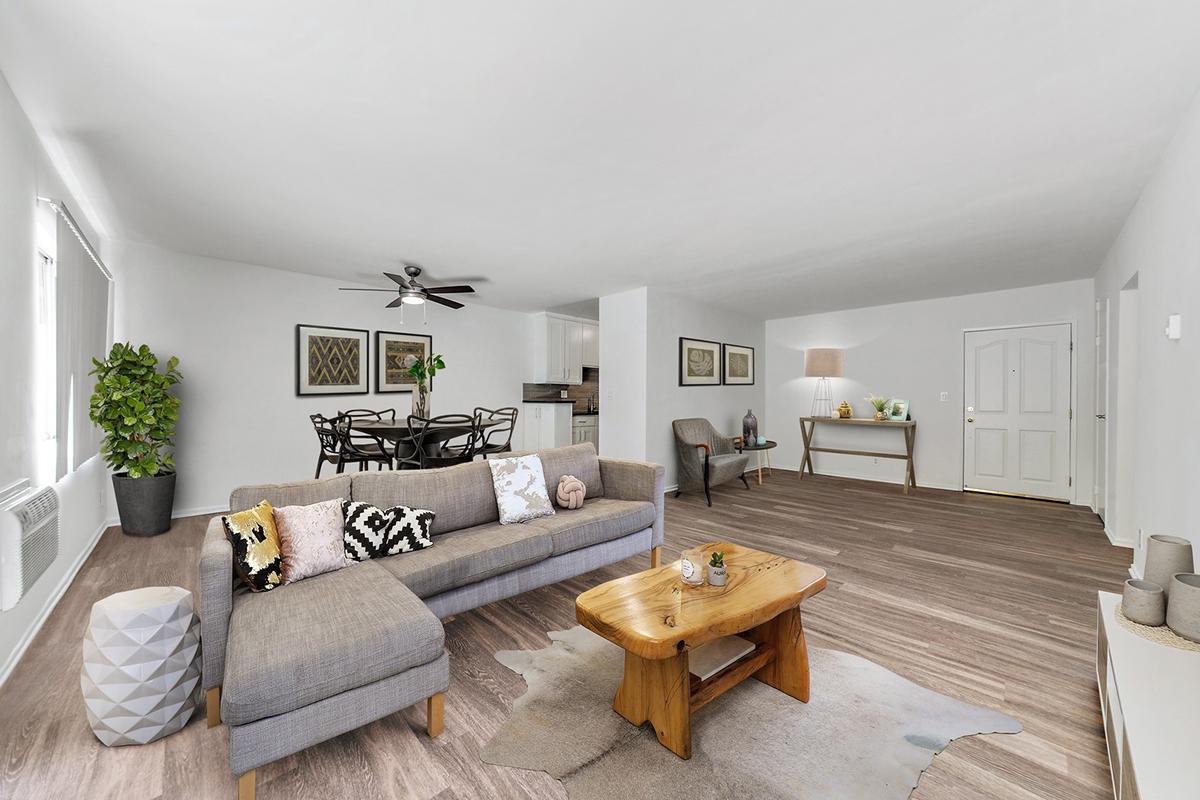 A modern living room featuring a gray sofa adorned with patterned cushions, a wooden coffee table, and a light-colored area rug. A potted plant adds greenery, while a dining area with a round table and chairs is visible in the background. Bright walls and a fan overhead create a spacious atmosphere.