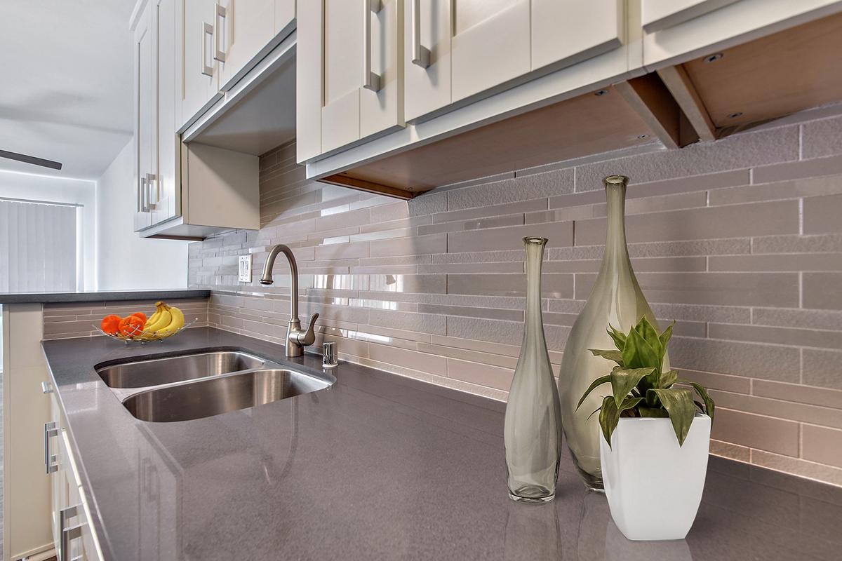 A modern kitchen countertop featuring a double sink, sleek cabinets, and a gray backsplash. Two glass vases and a small potted plant are displayed on the counter, alongside fresh fruits in a bowl. The overall design is clean and contemporary with a focus on light and neutral colors.
