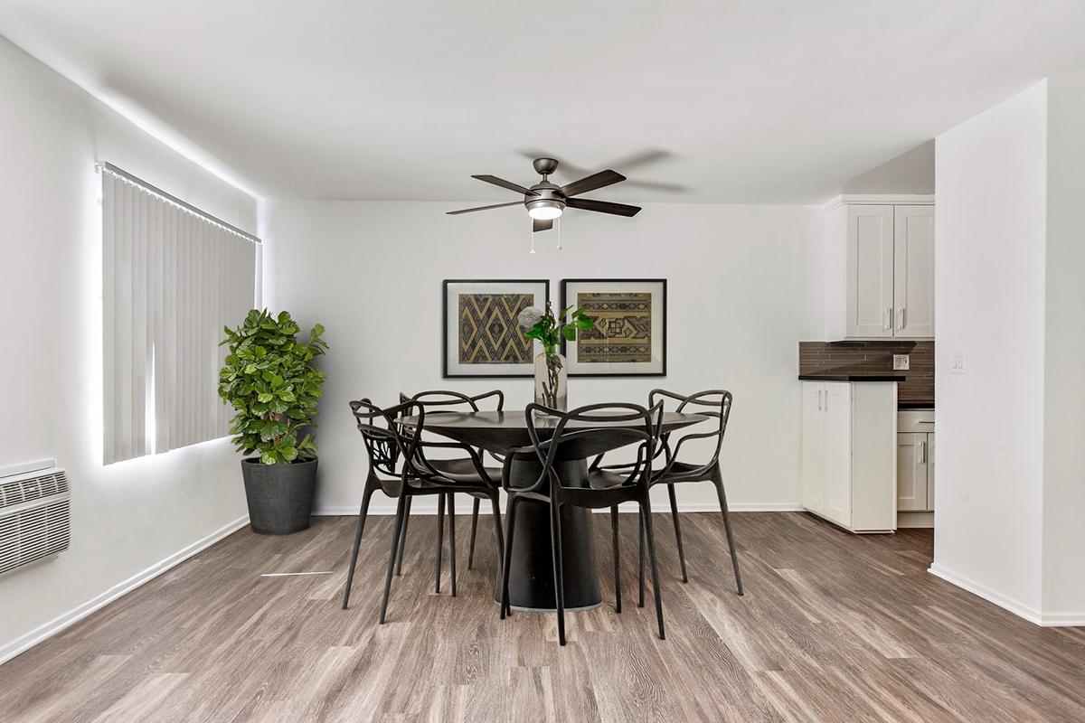 A modern dining area featuring a round black table surrounded by six black chairs. There is a potted plant in the corner and two framed artworks on the wall. The space has light-colored walls and a ceiling fan, with laminate flooring adding to the contemporary feel.