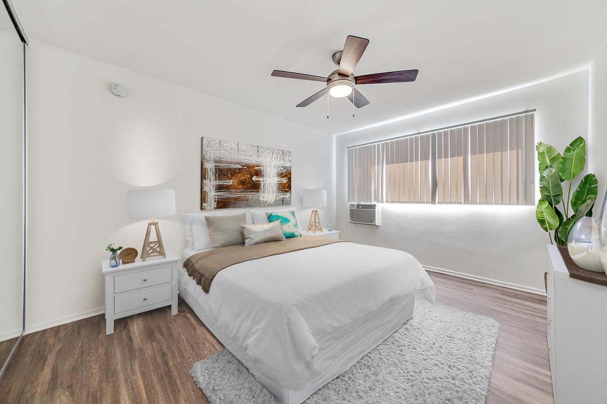 A bright and modern bedroom with a large bed dressed in white linens, accented by a brown throw. There are two bedside tables with lamps, a large piece of art on the wall, and a window with blinds letting in natural light. An air conditioning unit is visible, along with a potted plant in the corner. The floor is covered in a soft area rug.