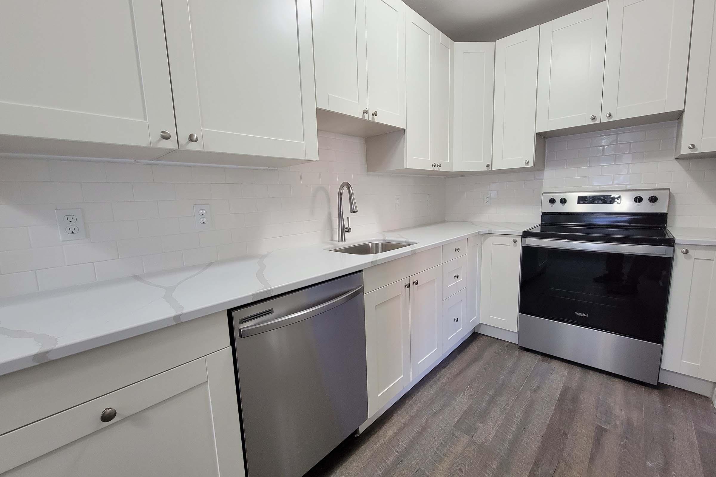Modern kitchen featuring white cabinetry, a stainless steel dishwasher, and an electric stove. The countertops are made of light-colored stone, and there’s a sleek sink with a modern faucet. The walls are tiled with white subway tiles, and the flooring is a dark wood finish, creating a contrast with the light cabinetry.
