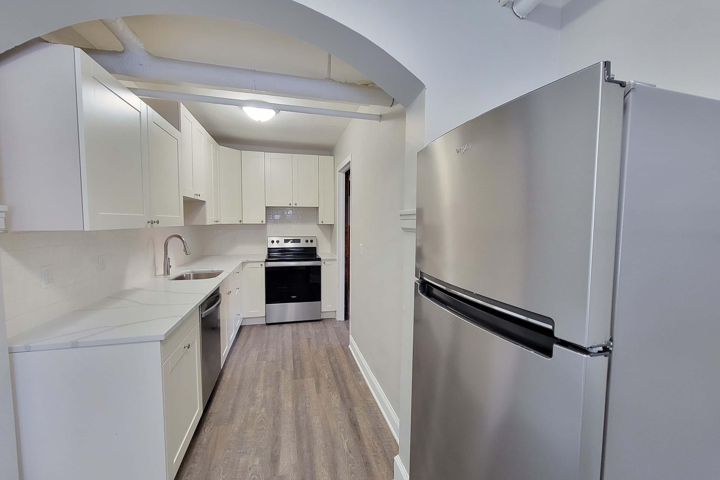 A modern kitchen featuring white cabinets, a stainless steel refrigerator, an oven, and a sink. The space has a light and airy feel with gray flooring and bright overhead lighting, showcased in an arched entryway.
