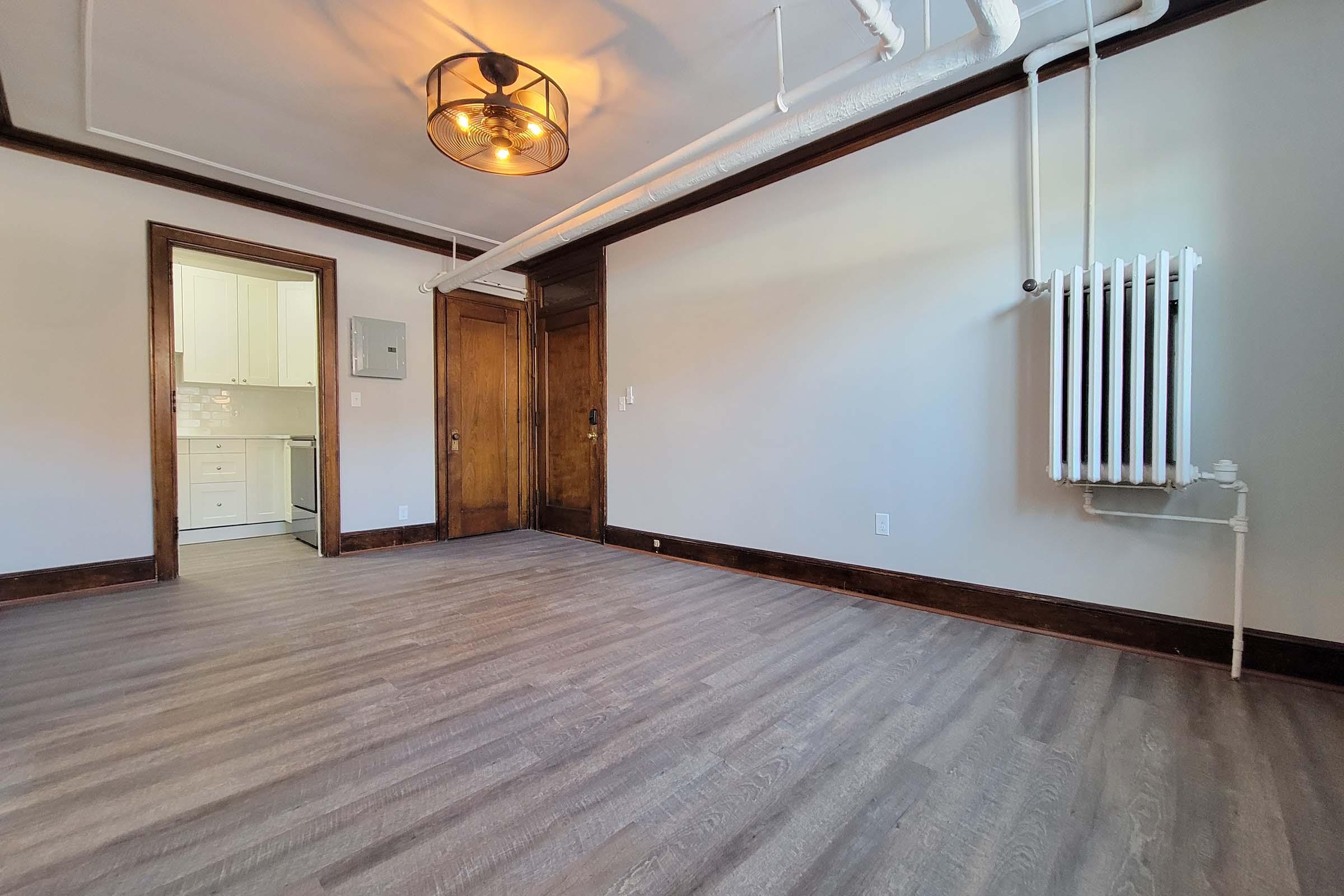 Interior of a spacious room featuring hardwood floors, a decorative ceiling light fixture, and a vintage radiator. A doorway leads to a kitchen area visible in the background. Neutral-colored walls complement the wood trim, creating a cozy atmosphere.