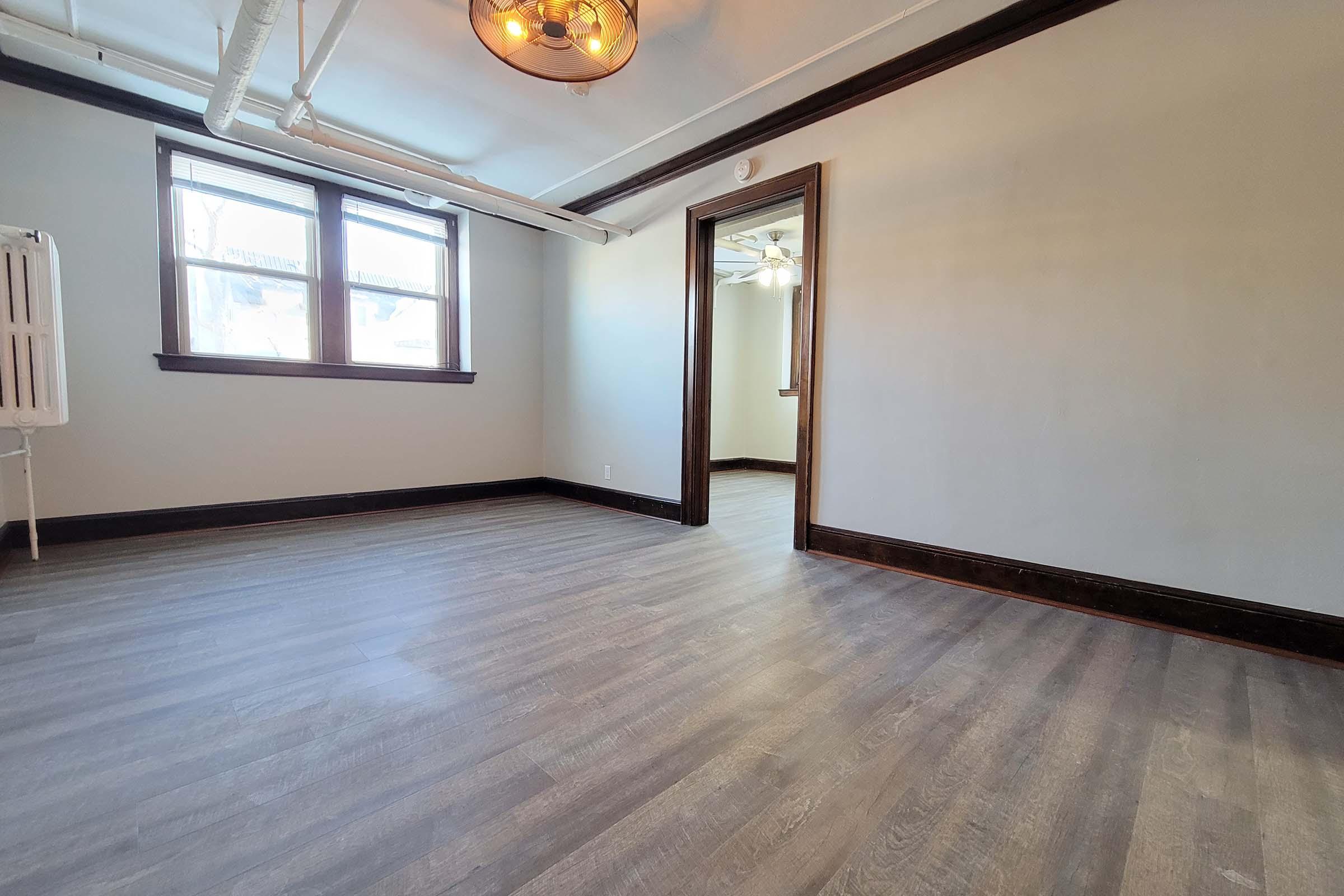 Empty room featuring light-colored walls, dark wood trim, and large windows. The floor is covered with laminate flooring, and there is a ceiling light fixture visible. A door leads to another room, and a radiator is placed against one wall. Natural light illuminates the space.