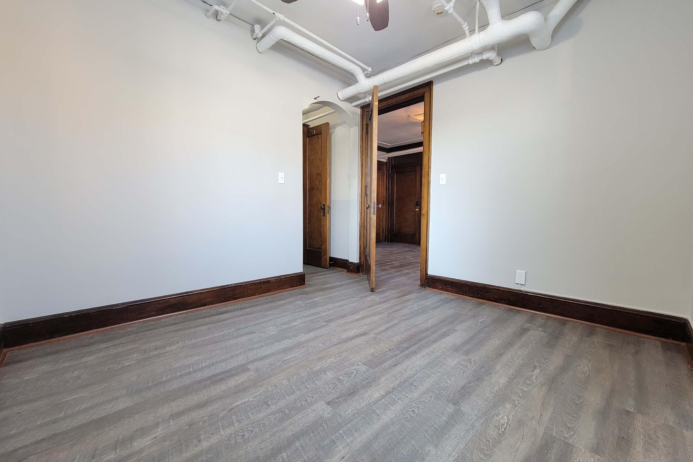 Empty room with light gray walls and wooden trim, featuring a door leading to another room. The floor is covered in light-colored laminate. Exposed pipes are visible on the ceiling. Natural light comes from an unseen source, giving the space a bright appearance.