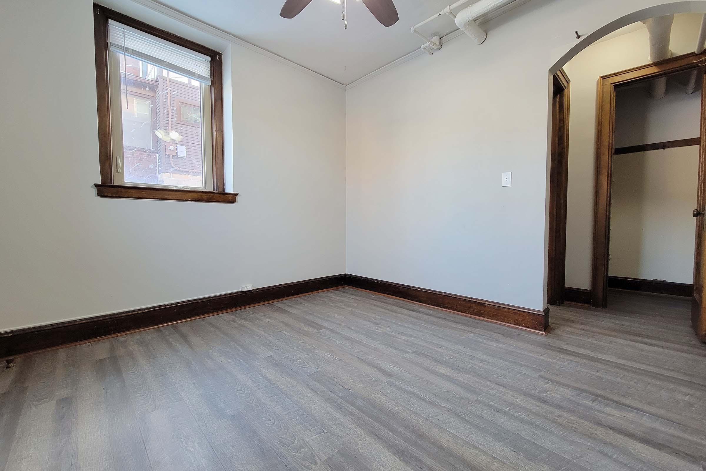 Empty room with light gray walls and a wooden floor. A ceiling fan is visible above, and there is a window letting in natural light. On one side, a doorway leads to another room, while the opposite wall features a small window. The space is uncluttered and ready for furniture.