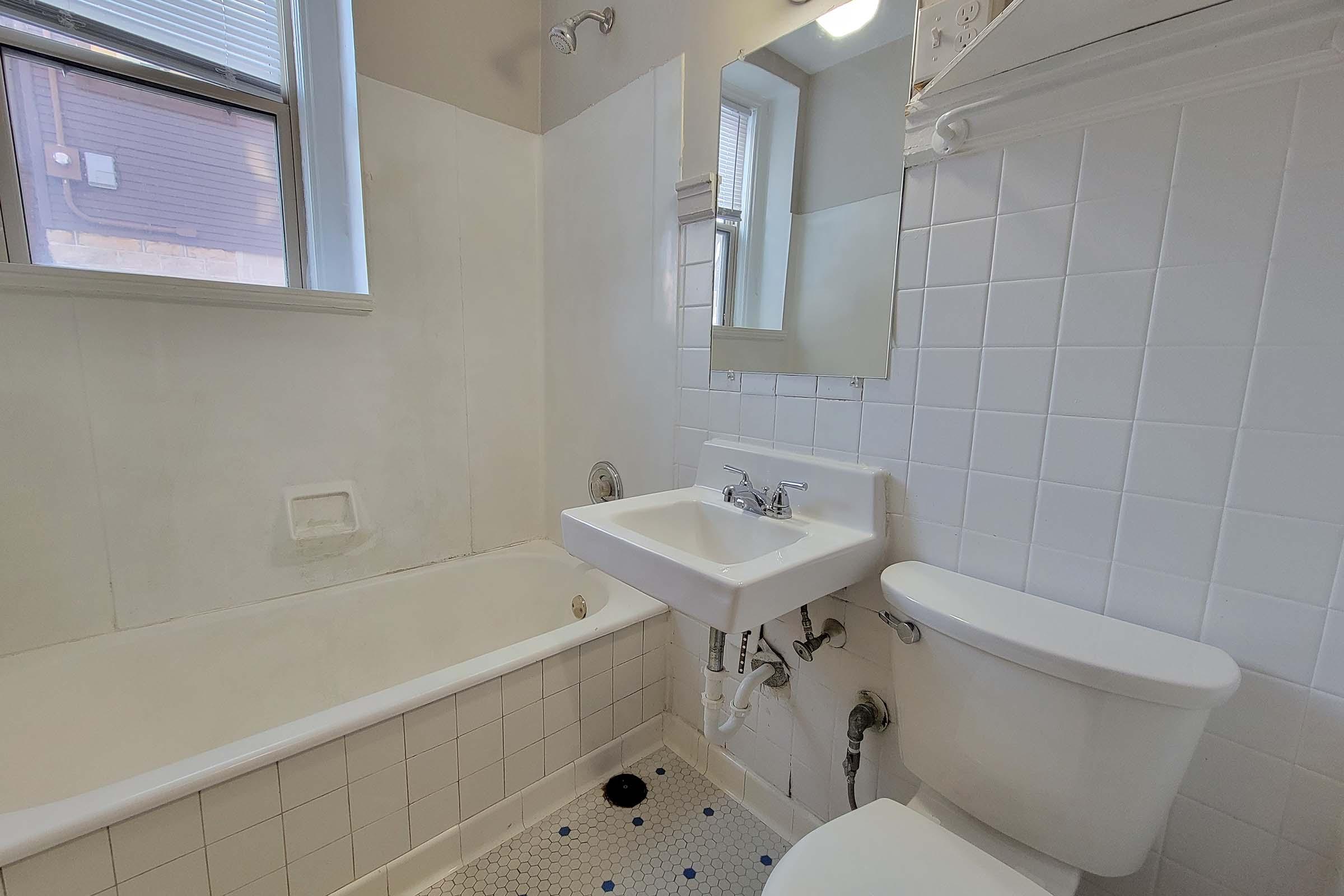 A small, clean bathroom featuring a white bathtub, a sink with a mirror above, and a toilet. The walls are tiled in white, and the floor has small hexagonal tiles. Natural light enters through a window, illuminating the space.