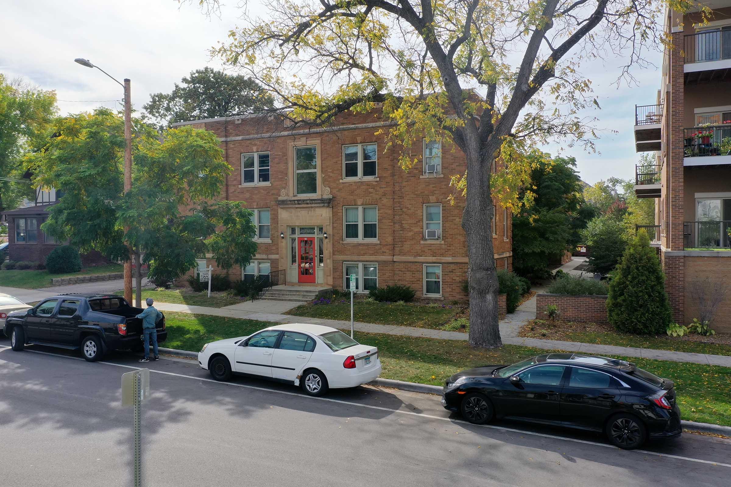 A view of a residential building with brick exterior and large windows. There are several parked cars in front, including a black truck, a white sedan, and a dark vehicle. A tree provides shade on the sidewalk, and there are neighboring buildings visible in the background. The scene is set in a suburban area with greenery.