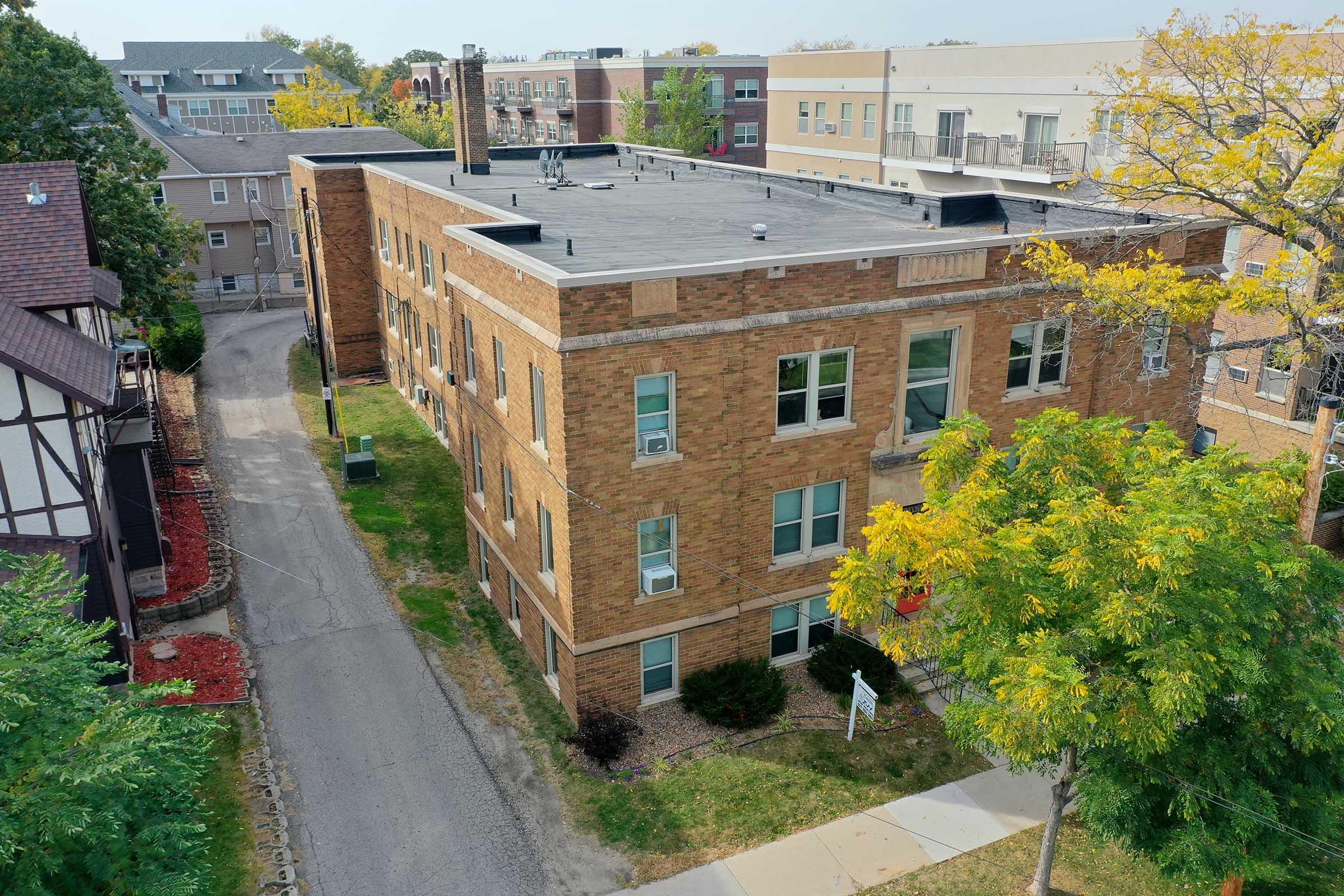 Aerial view of a brick apartment building surrounded by trees and grassy areas, with a paved road running alongside. In the background, additional residential buildings are visible, showcasing a suburban neighborhood atmosphere. The scene is bright and reflects a clear day.