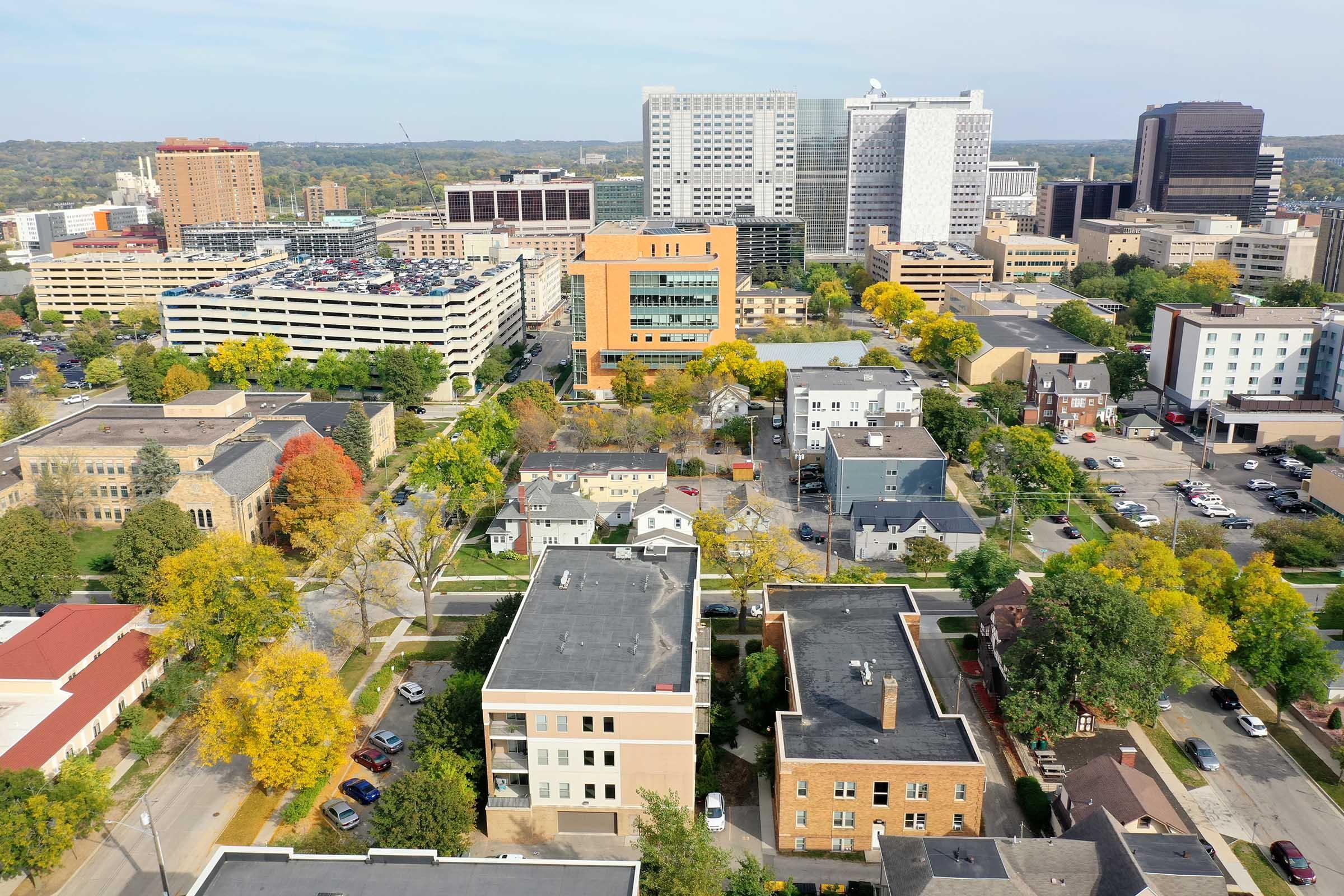 Aerial view of a cityscape featuring a mix of modern buildings and residential homes. Lush green trees with autumn foliage are visible, contrasting with concrete structures. In the background, tall skyscrapers and commercial buildings dominate the skyline, showcasing urban development.