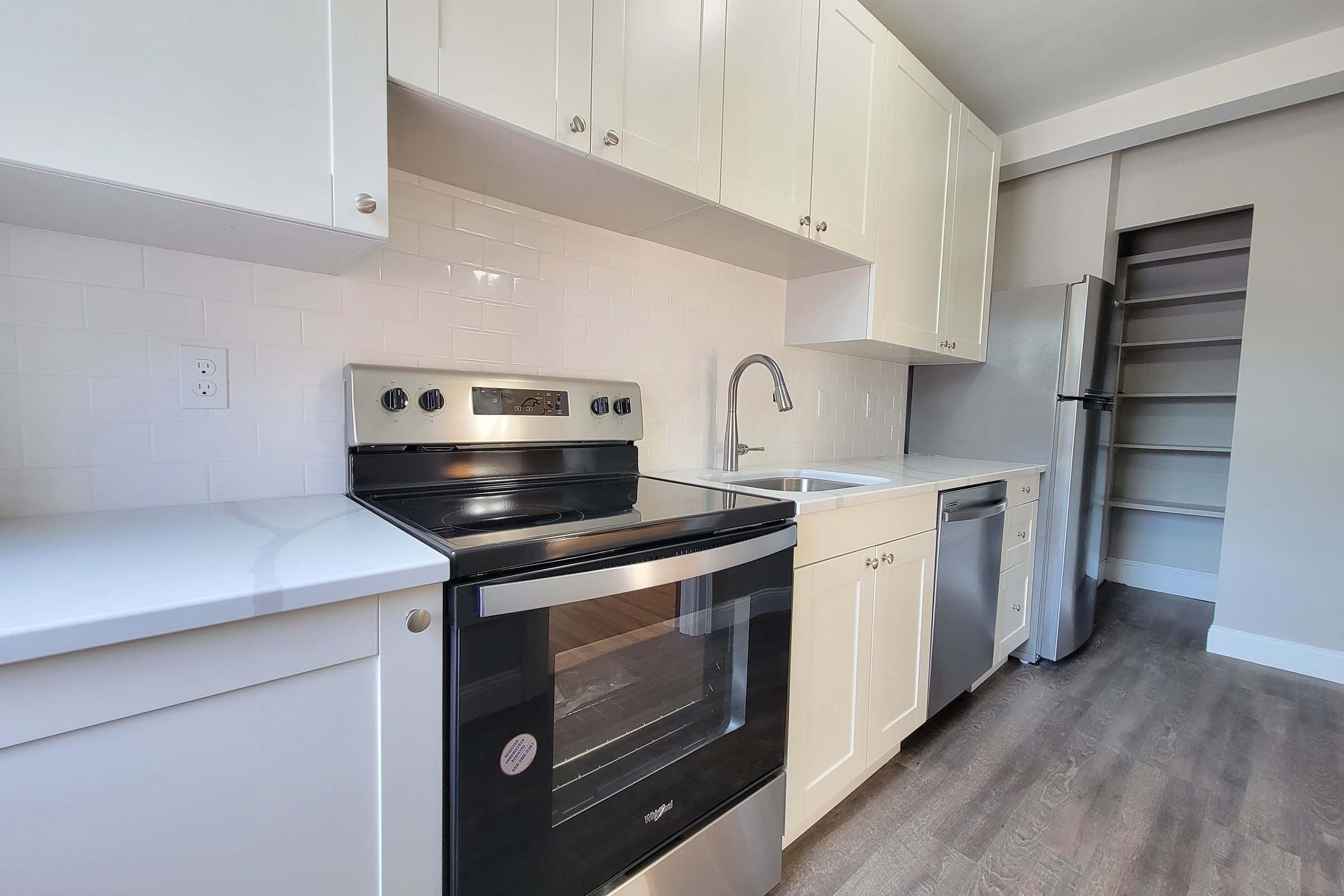 A modern kitchen featuring white cabinetry, an electric stove, a sink with a faucet, a dishwasher, and a stainless steel refrigerator. The countertops are light-colored, and the floor has a wood-like finish. There is a pantry or storage area visible in the background.
