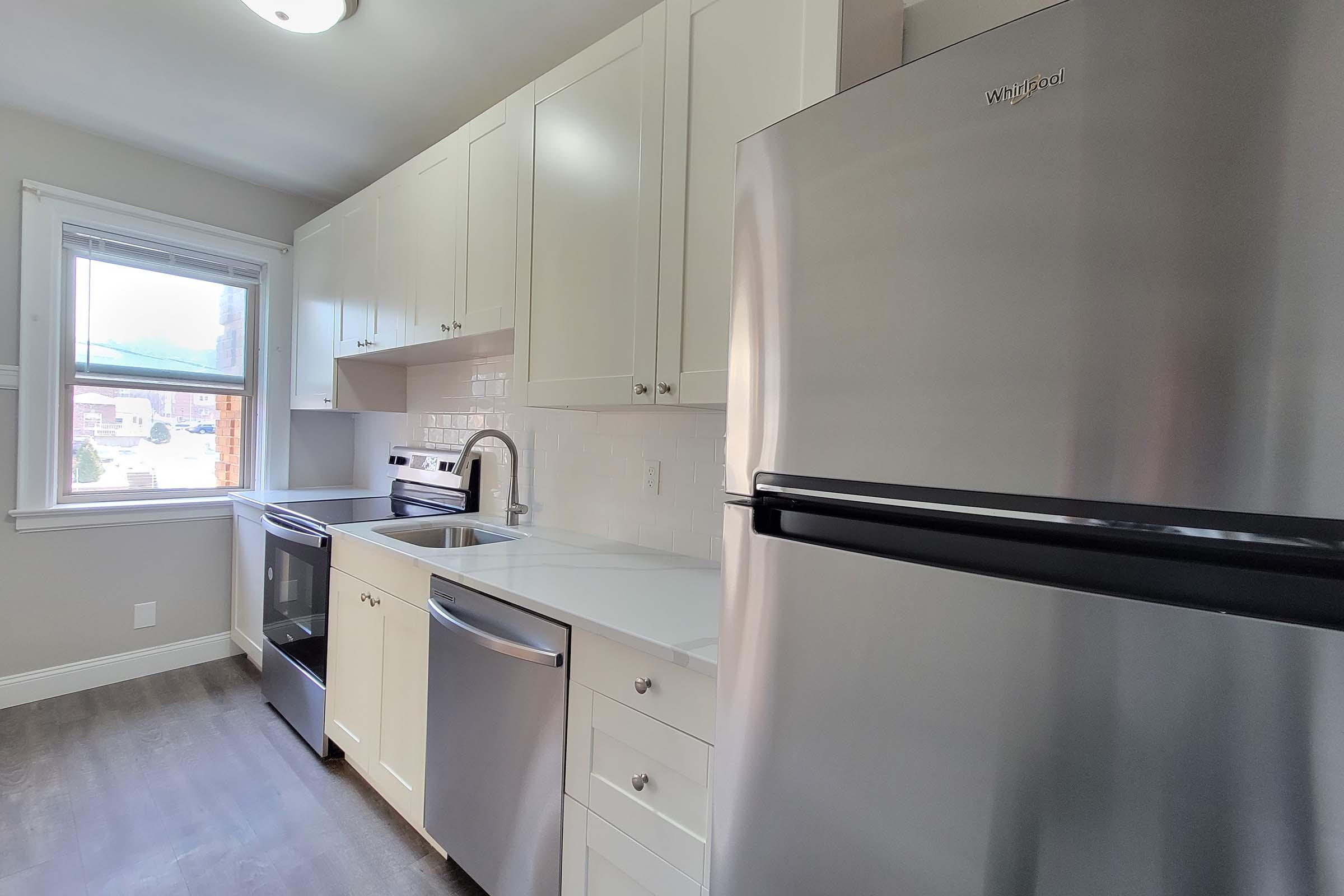 A modern kitchen featuring white cabinets, a stainless steel refrigerator, a sink, and a gas stove. The countertop is light-colored, and a window offers natural light. A dishwasher is also included, and the flooring is a dark wood tone.