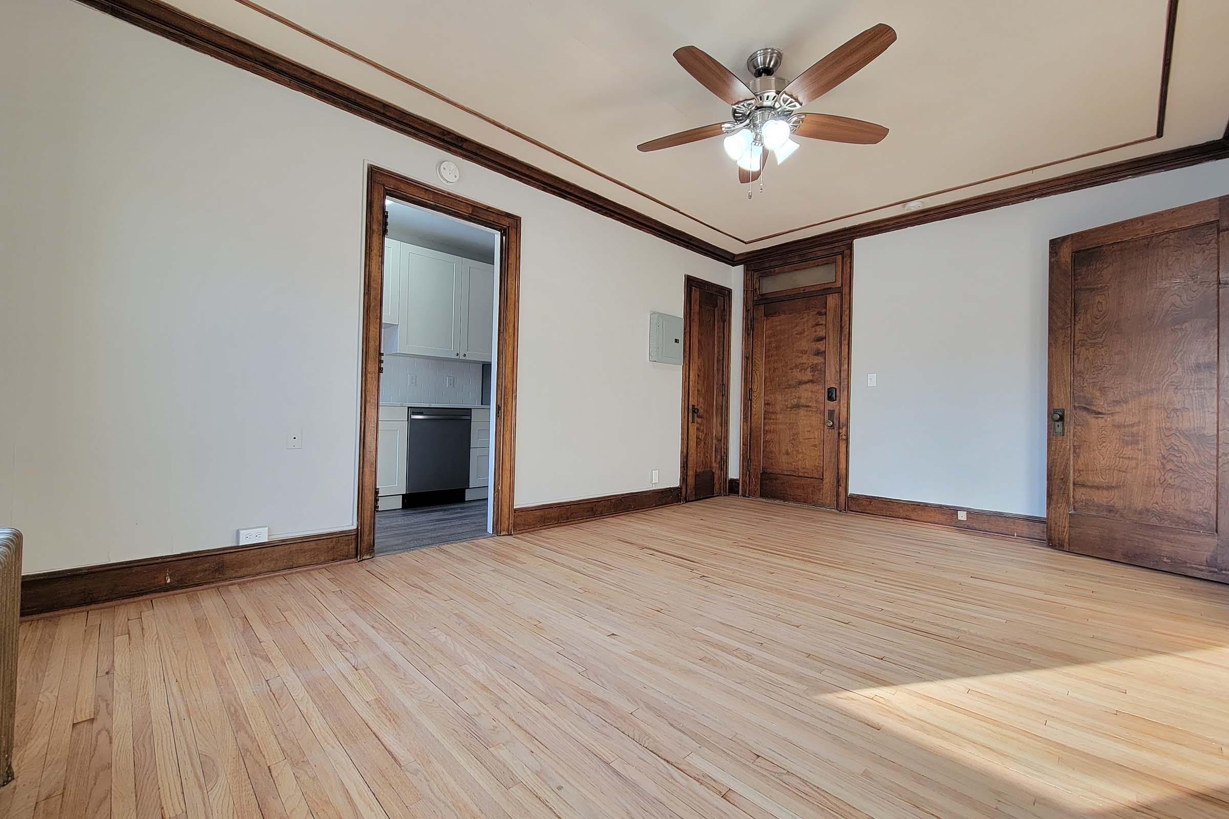 A spacious, empty room with hardwood floors and a ceiling fan. Light streams in through an open doorway leading to a kitchen. The walls are painted light gray, and there are wooden trim details around the doors. A closet or storage area is visible in the back wall, enhancing the room's functionality.