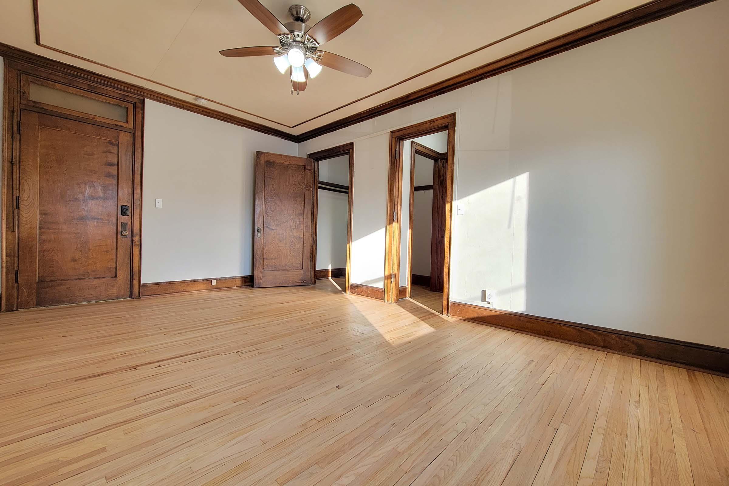 A spacious, vacant room featuring hardwood flooring, a ceiling fan, and wooden trim. Two doors lead to adjacent spaces, with sunlight streaming in through a window, creating a warm atmosphere. The walls are painted a light color, enhancing the room's openness.