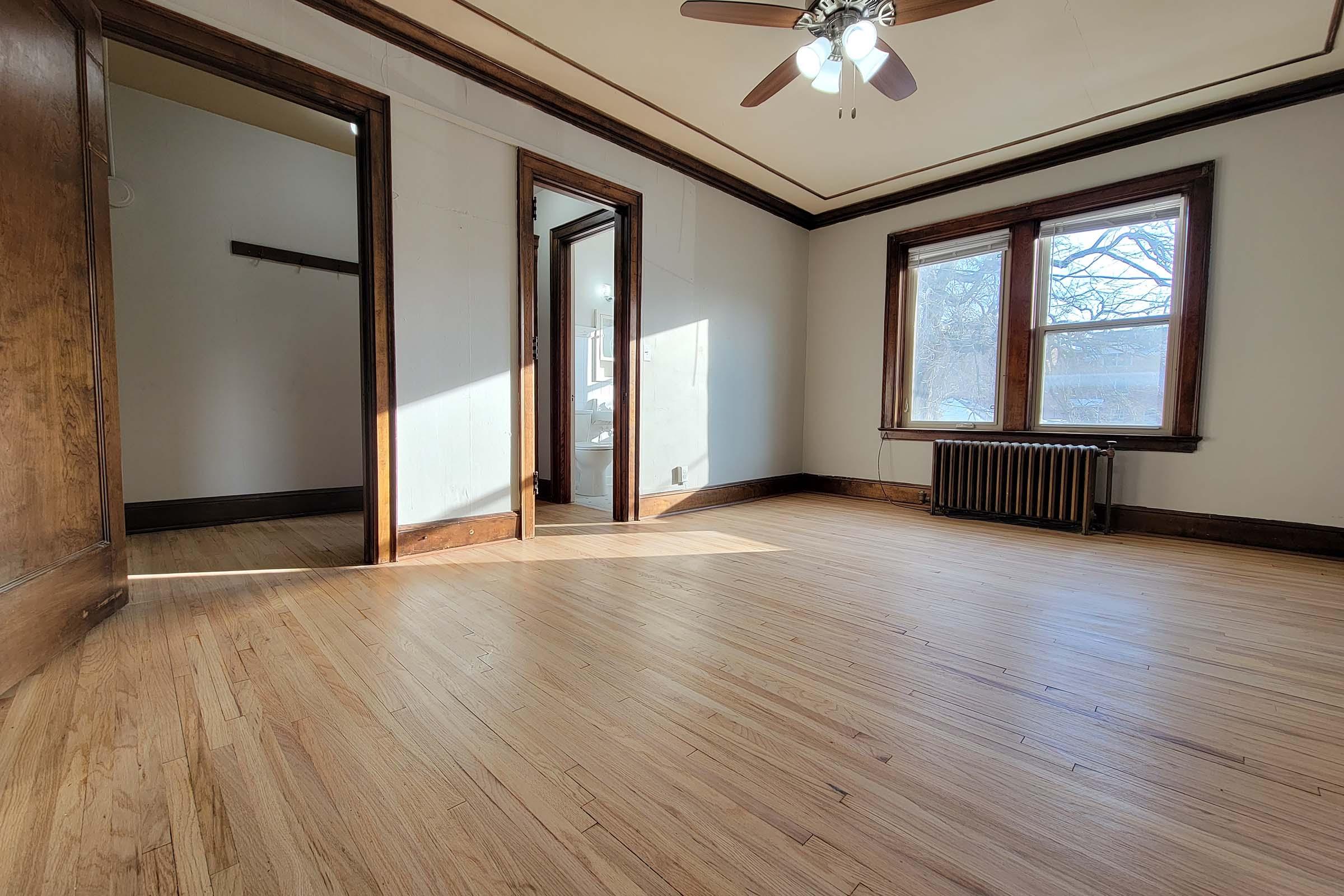 Empty room with hardwood floors, featuring large windows allowing natural light. There are wooden trim details around the windows and doors. A small radiator is visible along the wall, and a closet opening is seen in the background. The overall setting is spacious and well-lit.