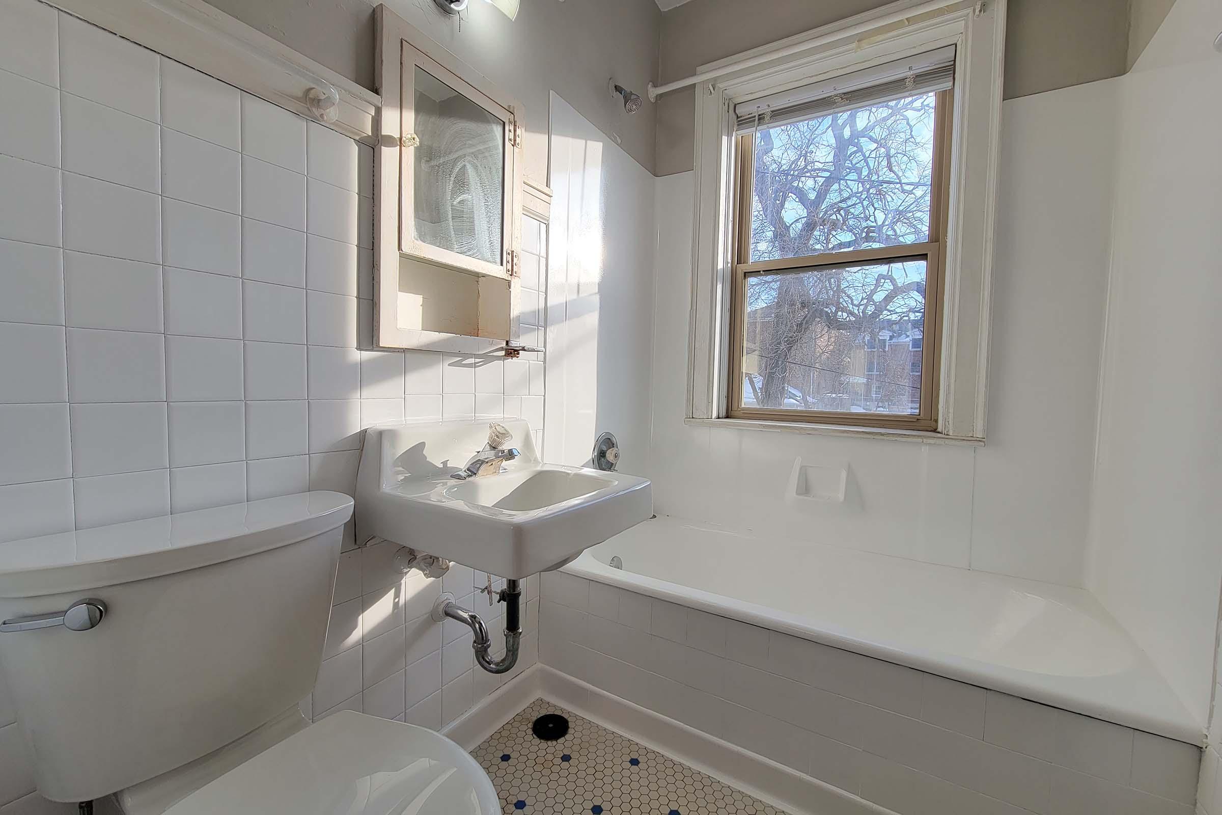 A bright, clean bathroom featuring a bathtub, a white sink mounted on the wall, and a toilet. Natural light streams in from a window with a tree visible outside. The walls are tiled with white tiles, and the floor has hexagonal black and white tiles. A mirror is mounted above the sink.
