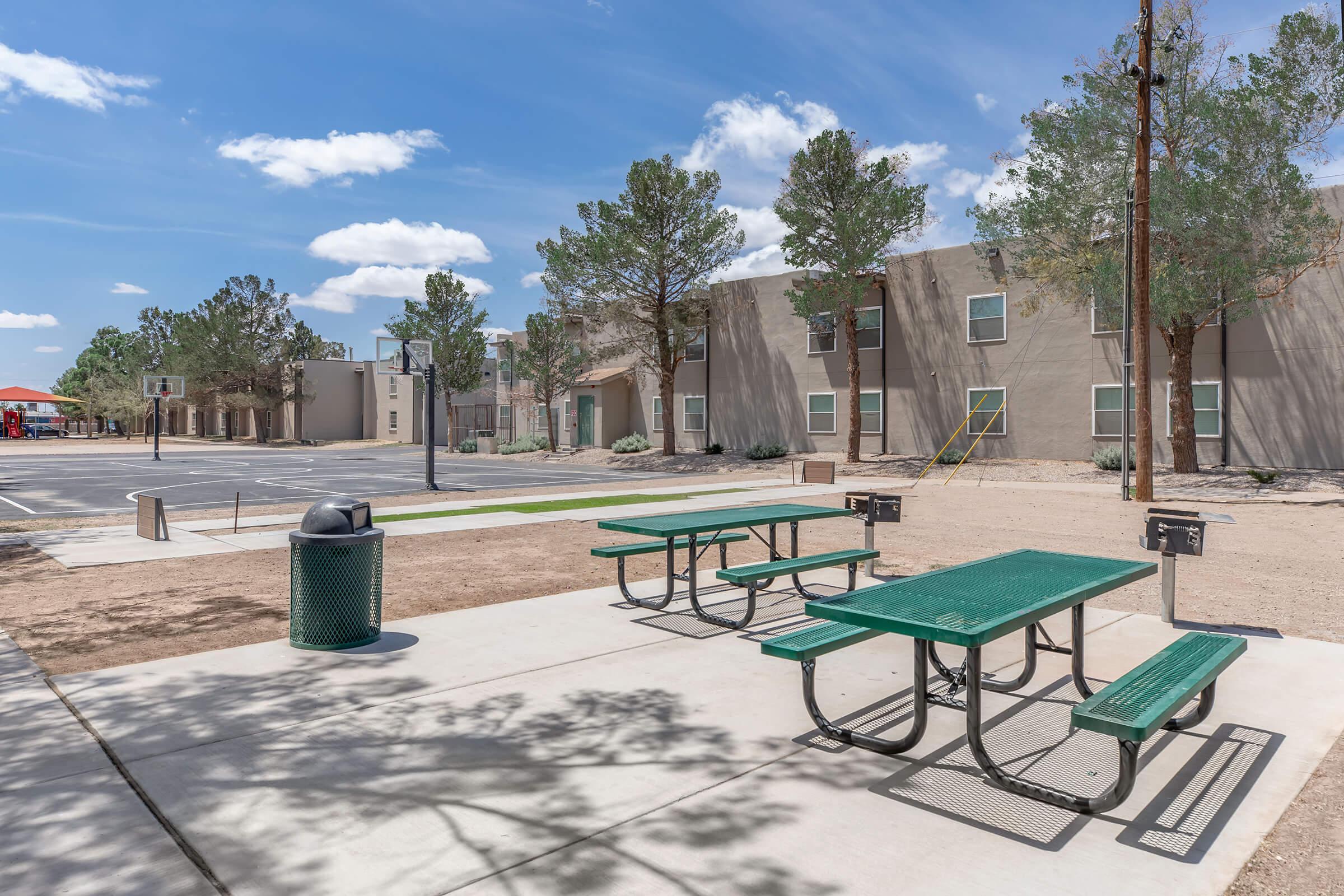 A view of a public area featuring green picnic tables and a trash can on a concrete surface. Surrounding the area are sparse trees and a beige building in the background under a blue sky with clouds. The space is clean and well-maintained, suitable for outdoor gatherings or leisure activities.