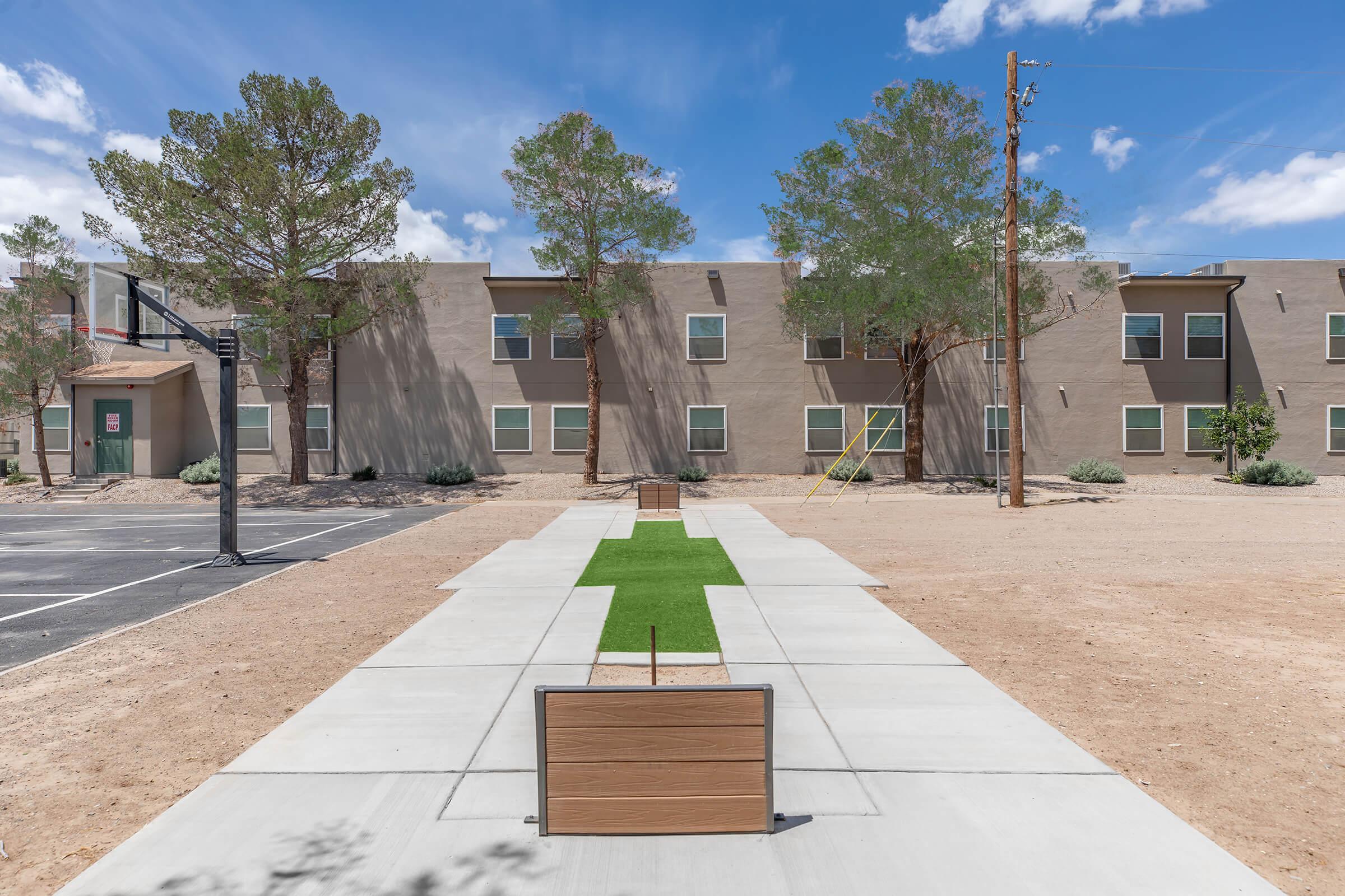 A pathway featuring green grass sections leads toward a series of residential buildings in a desert setting. Nearby, a basketball hoop is visible, along with sparse landscaping, utility poles, and a clear blue sky. The area appears well-maintained but relatively empty.