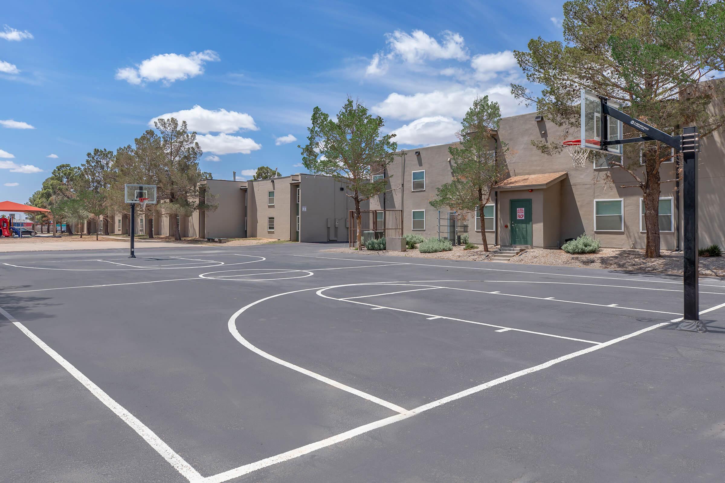 Empty outdoor basketball court with two hoops, surrounded by trees and residential buildings. The court features white lines on a dark surface, with blue sky and fluffy clouds visible overhead. In the background, a playground structure is partially visible.