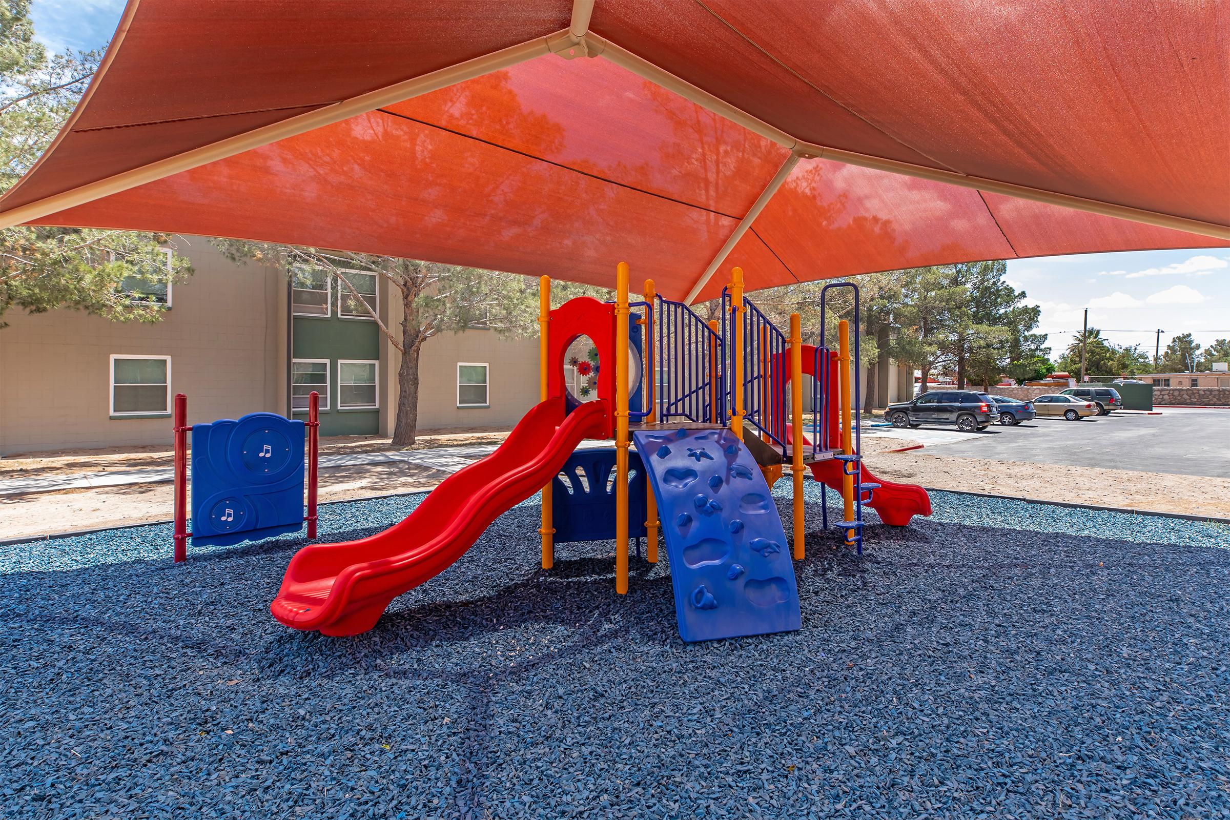 Colorful playground equipment featuring a red slide, climbing wall, and various structures, all covered by a large shade canopy. The playground is surrounded by a blue rubber surface and is located near residential buildings with parked cars in the background.