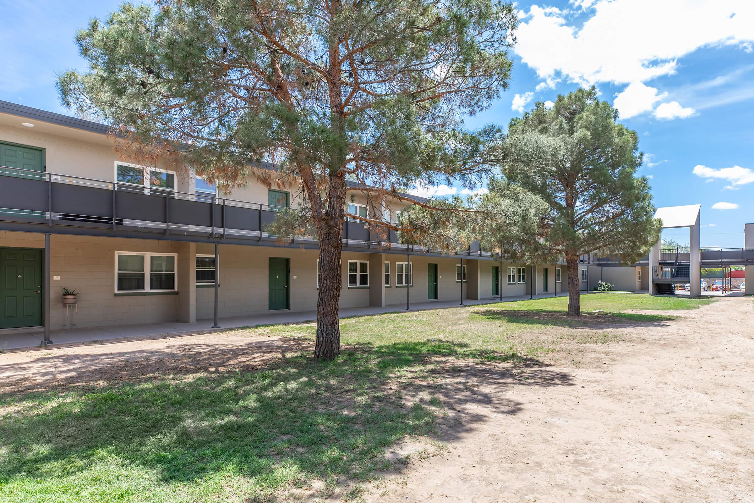 A low-rise building with multiple entrances, surrounded by green grass and trees. The building features balconies along the upper level and is situated under a clear blue sky with scattered clouds. The area appears well-maintained, providing a welcoming atmosphere.