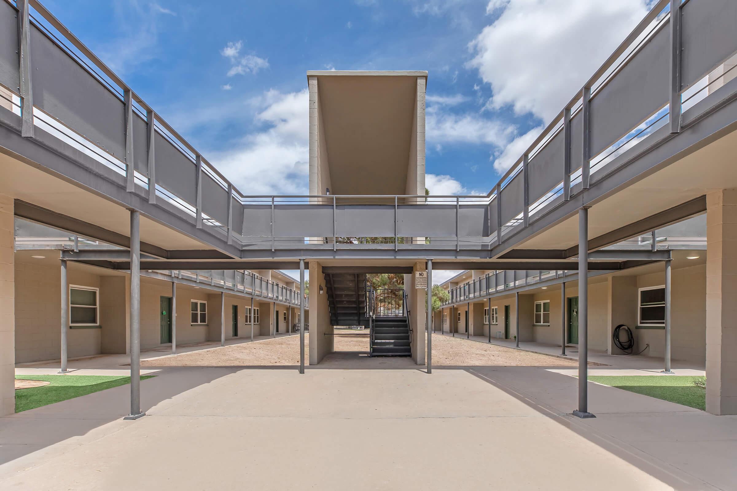 A view of a modern courtyard in a multi-level building with rectangular balconies and an upper shaded area. The pathway leads to a central staircase, flanked by symmetrical rows of doorways. The scene is bright, featuring blue skies with scattered clouds and touches of greenery.