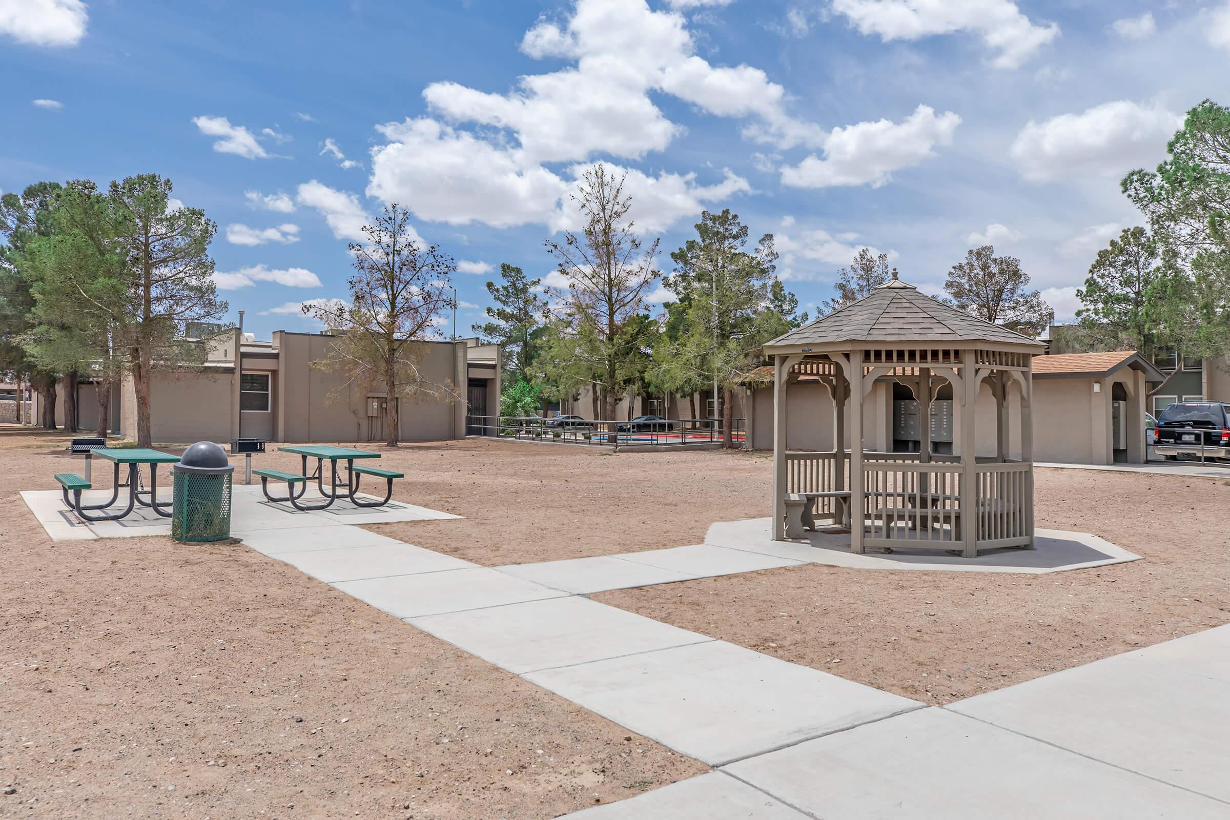 A landscaped area featuring a gazebo set on a concrete pathway. Nearby, there are picnic tables and a garbage bin. Surrounding trees provide some shade, and buildings are visible in the background under a blue sky with fluffy clouds. The scene conveys a quiet outdoor space ideal for relaxation.