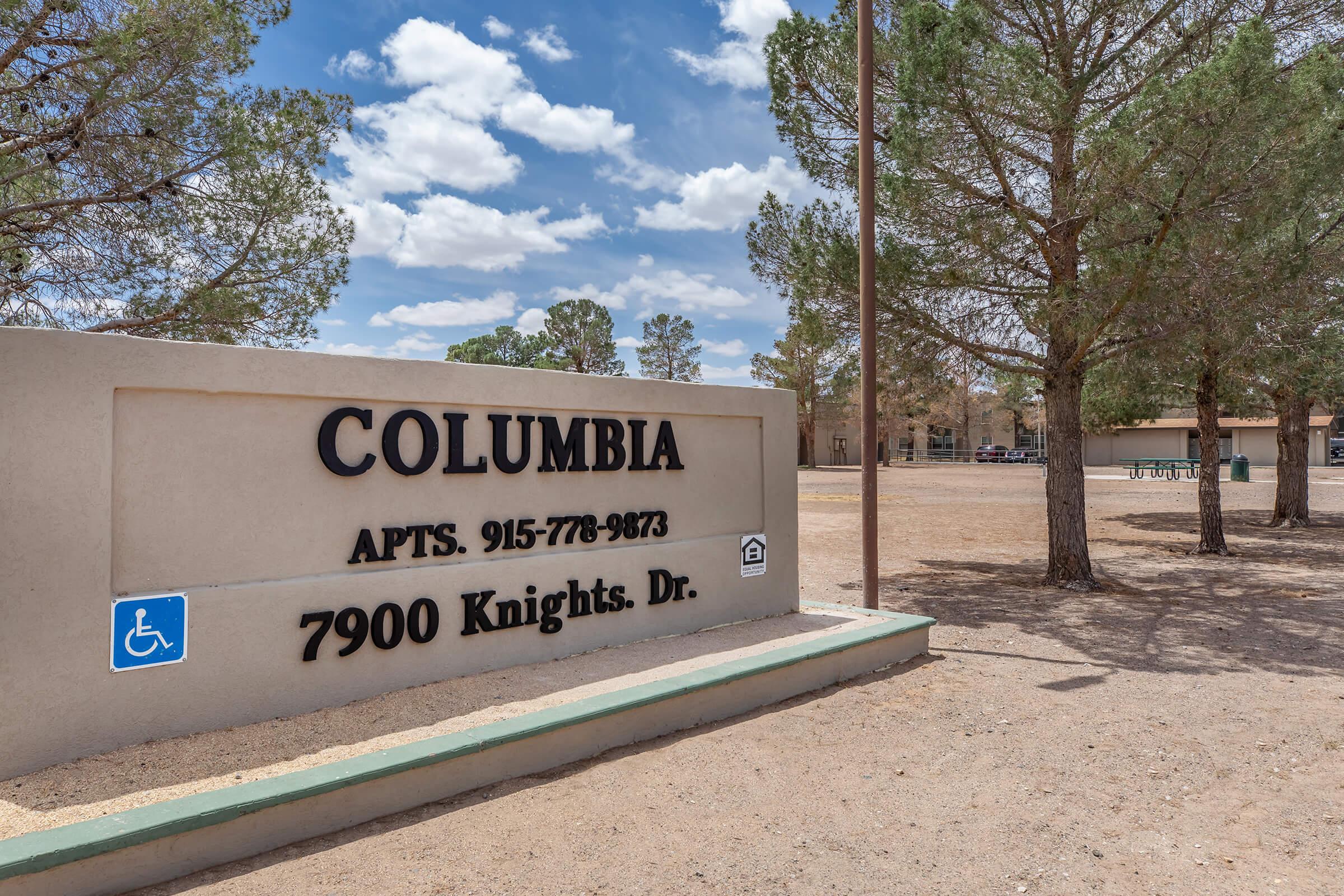 Sign for Columbia Apartments located at 7900 Knights Dr, featuring the phone number 915-778-9673. Surrounding area includes sparse vegetation and a parking lot. The sky is partly cloudy, providing a bright atmosphere.