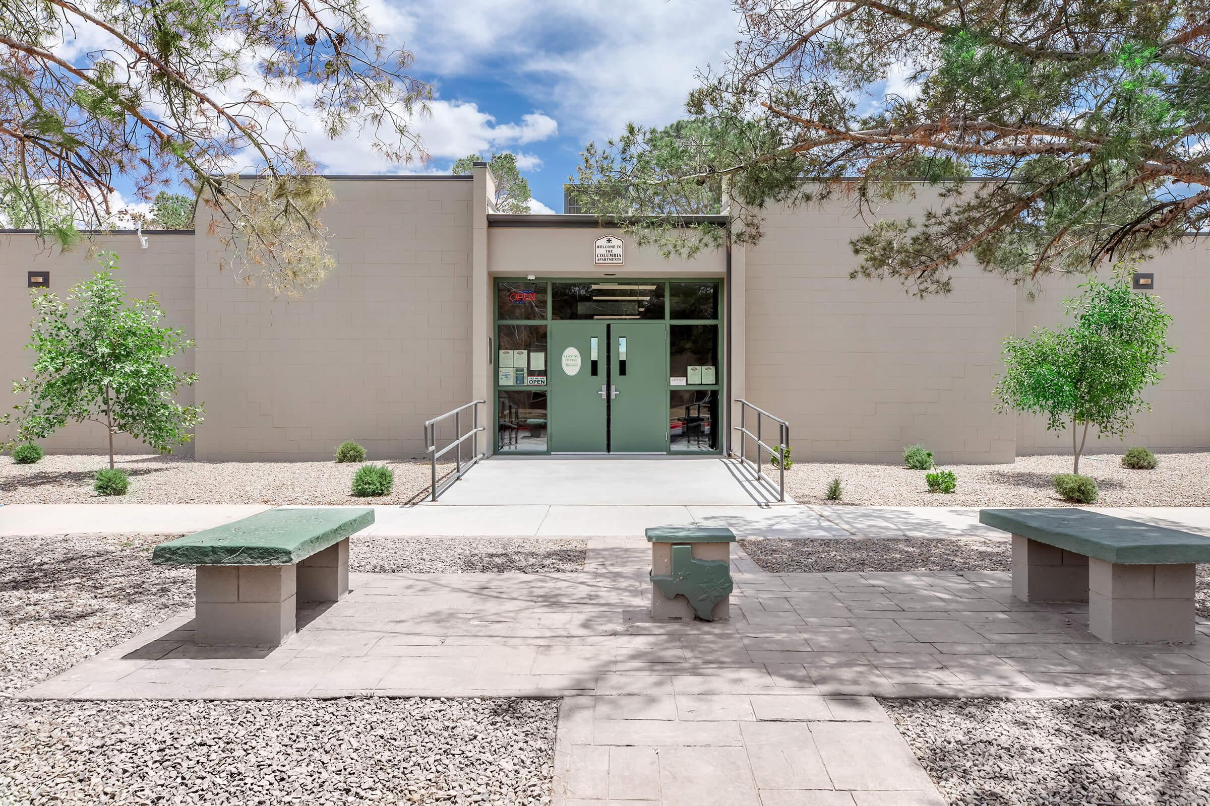 A modern building entrance with double green doors, surrounded by landscaped areas featuring small trees and gravel. There are concrete benches in front, and a clear blue sky with some clouds overhead. The setting appears inviting and well-maintained.