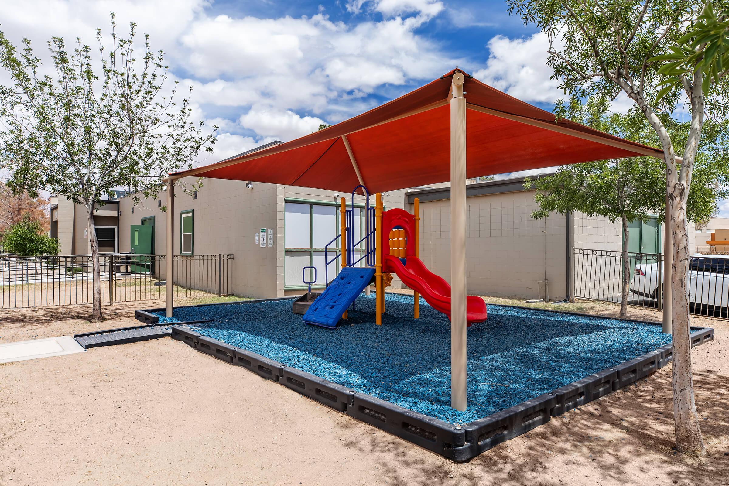 Colorful playground featuring a red slide and blue climbing structure under a shaded canopy. The play area is surrounded by low fencing, with trees visible in the background and a building in the distance. The ground is covered with blue rubber material for safety.