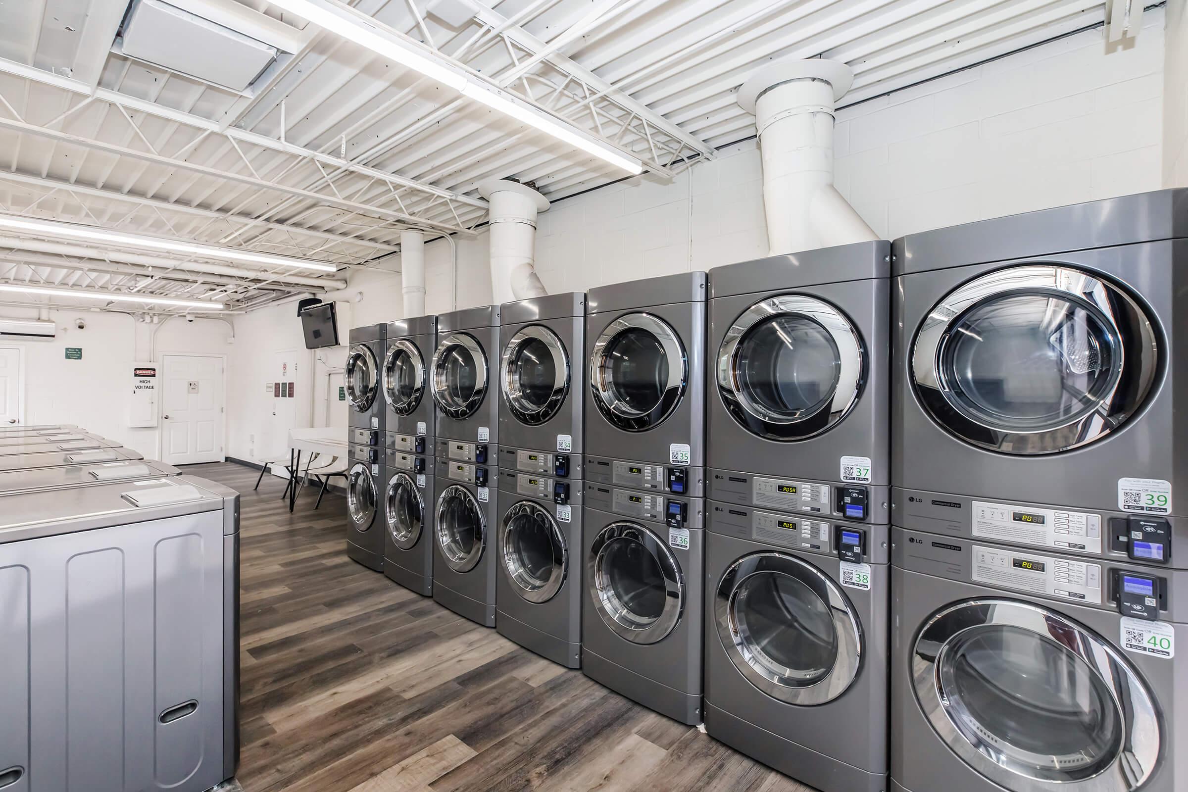 A modern laundry facility featuring a row of silver commercial washing machines and dryers against a white wall. The flooring is wood-like, and the space is well-lit with overhead lighting. There are folding tables and additional amenities visible in the background.