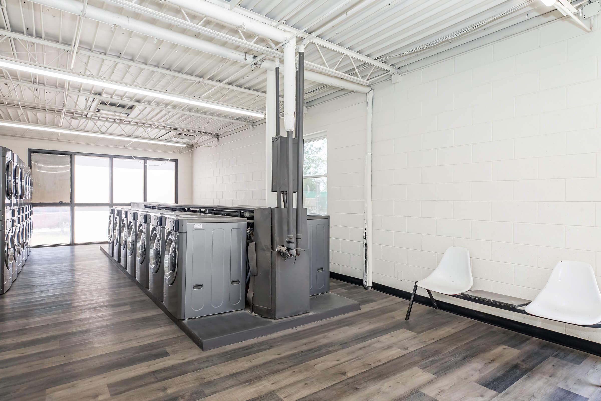 A spacious laundromat with several washing machines lined up against one wall. The interior has a modern design with white walls, large windows letting in natural light, and a bench for seating. The flooring is wooden, creating a clean and inviting atmosphere.