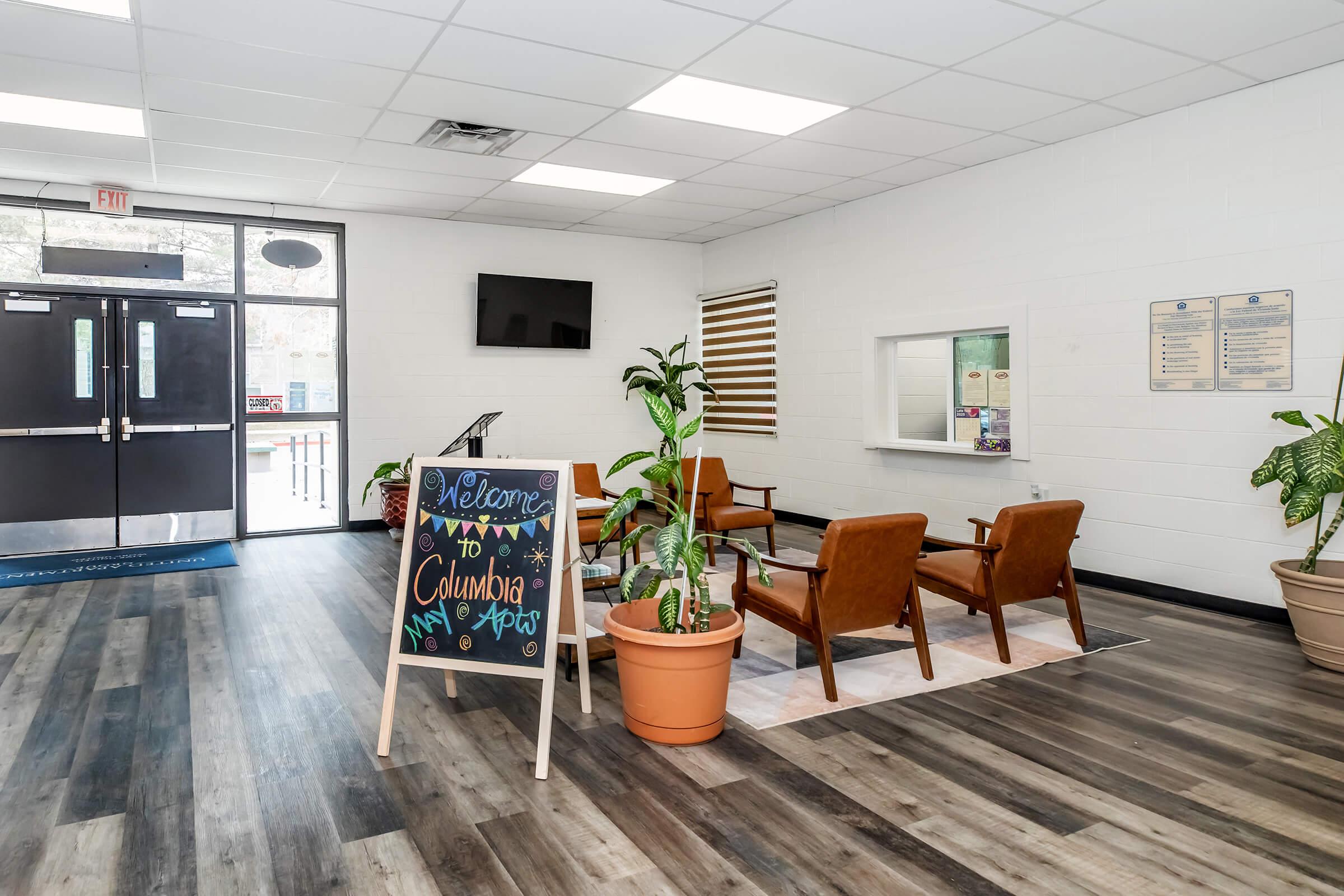A welcoming interior of a lobby featuring two brown chairs, a large potted plant, and a decorative sign that reads "Welcome to Columbia." The space has light-colored walls, a TV on the wall, and a reception area with informative posters. The flooring is dark wood, contributing to a modern look.