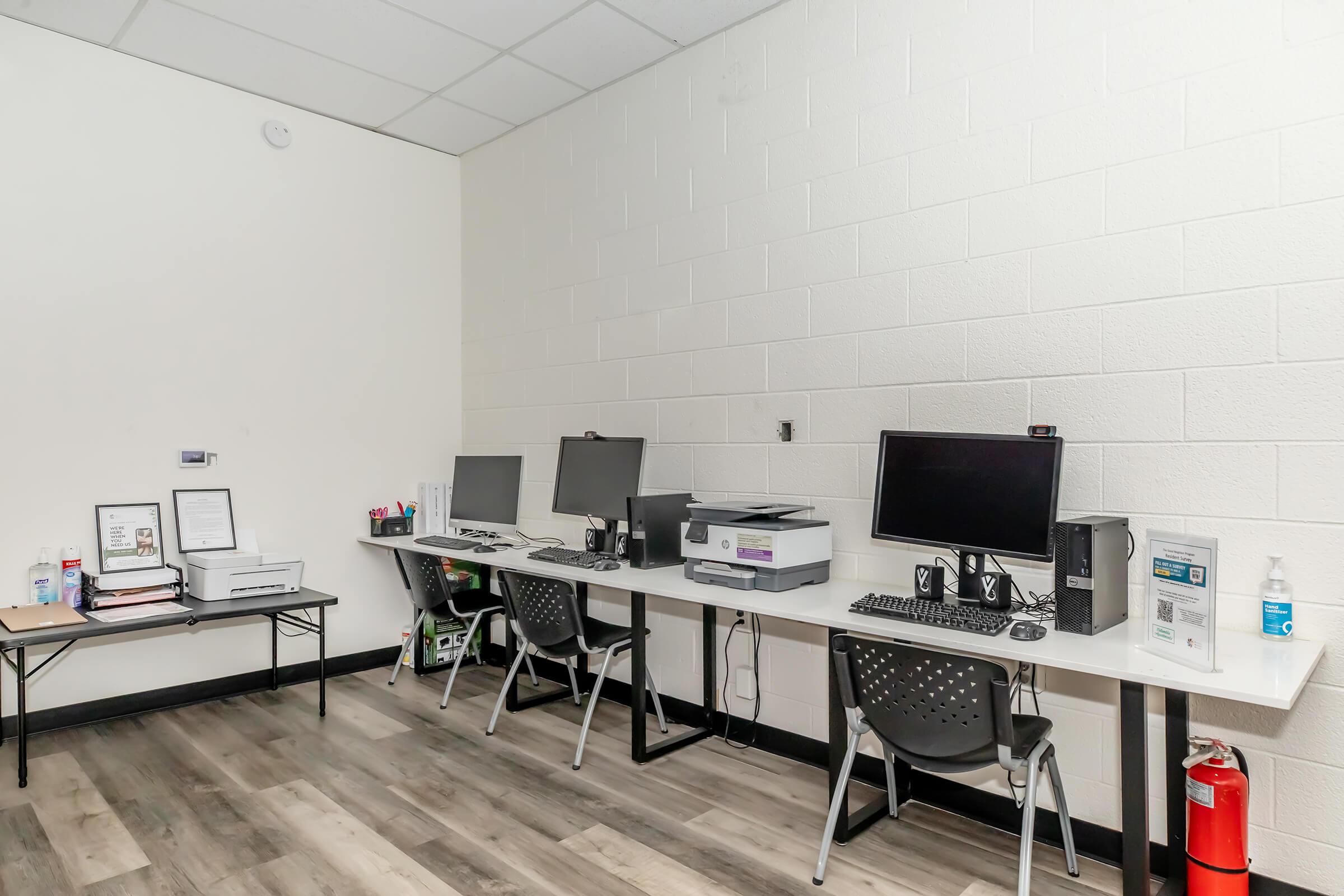 A minimalist office space featuring two desks with desktop computers and monitors. There's a printer on one desk, and the other has various office supplies. The room has light-colored walls and a wooden floor, creating a clean and organized environment.