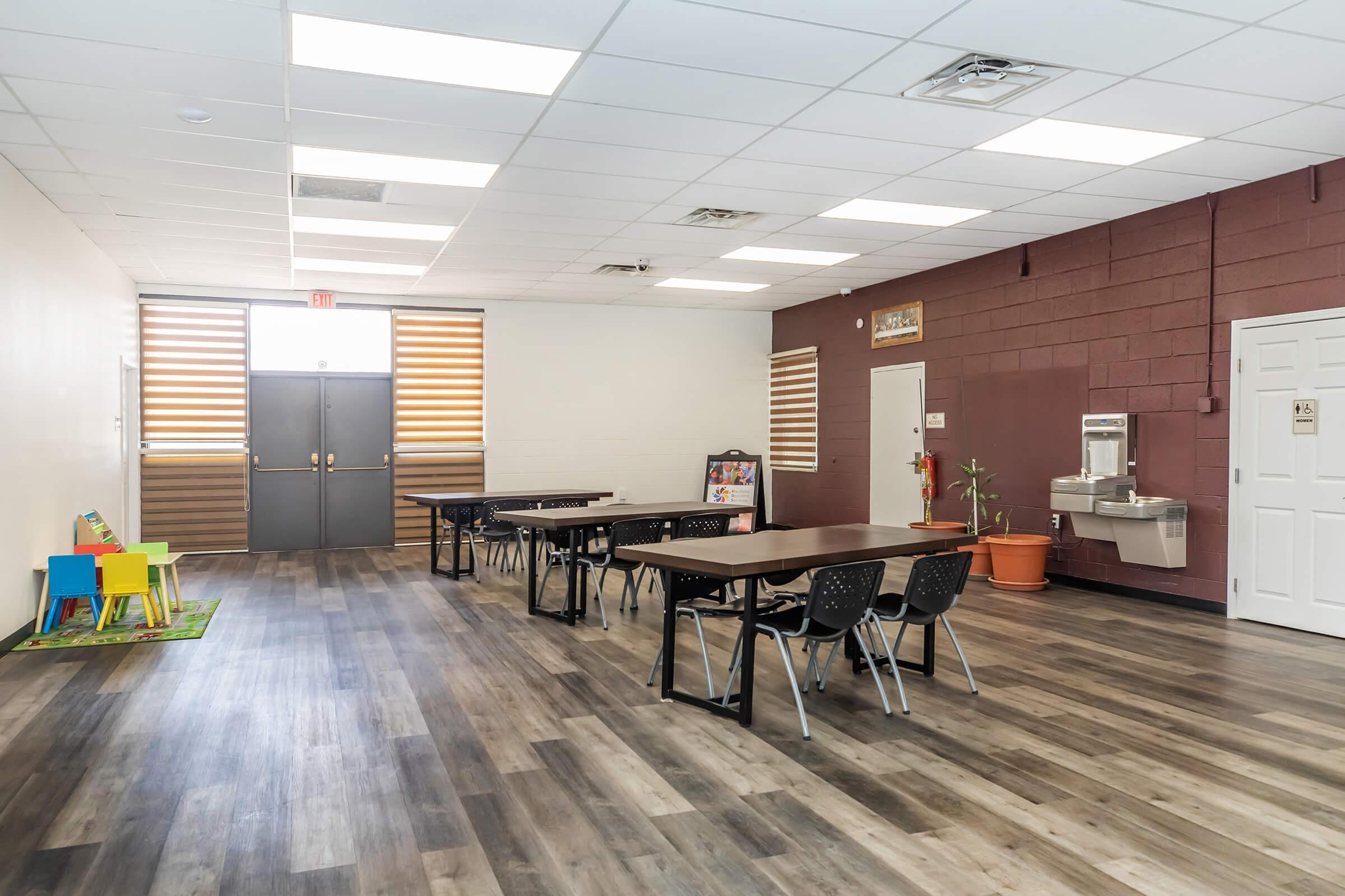 A spacious, well-lit room with a wooden floor, featuring multiple tables with black chairs. There's a water fountain along the wall and large windows with wooden blinds. A green rug with colorful chairs is visible in one corner, creating a welcoming atmosphere suitable for meetings or gatherings.