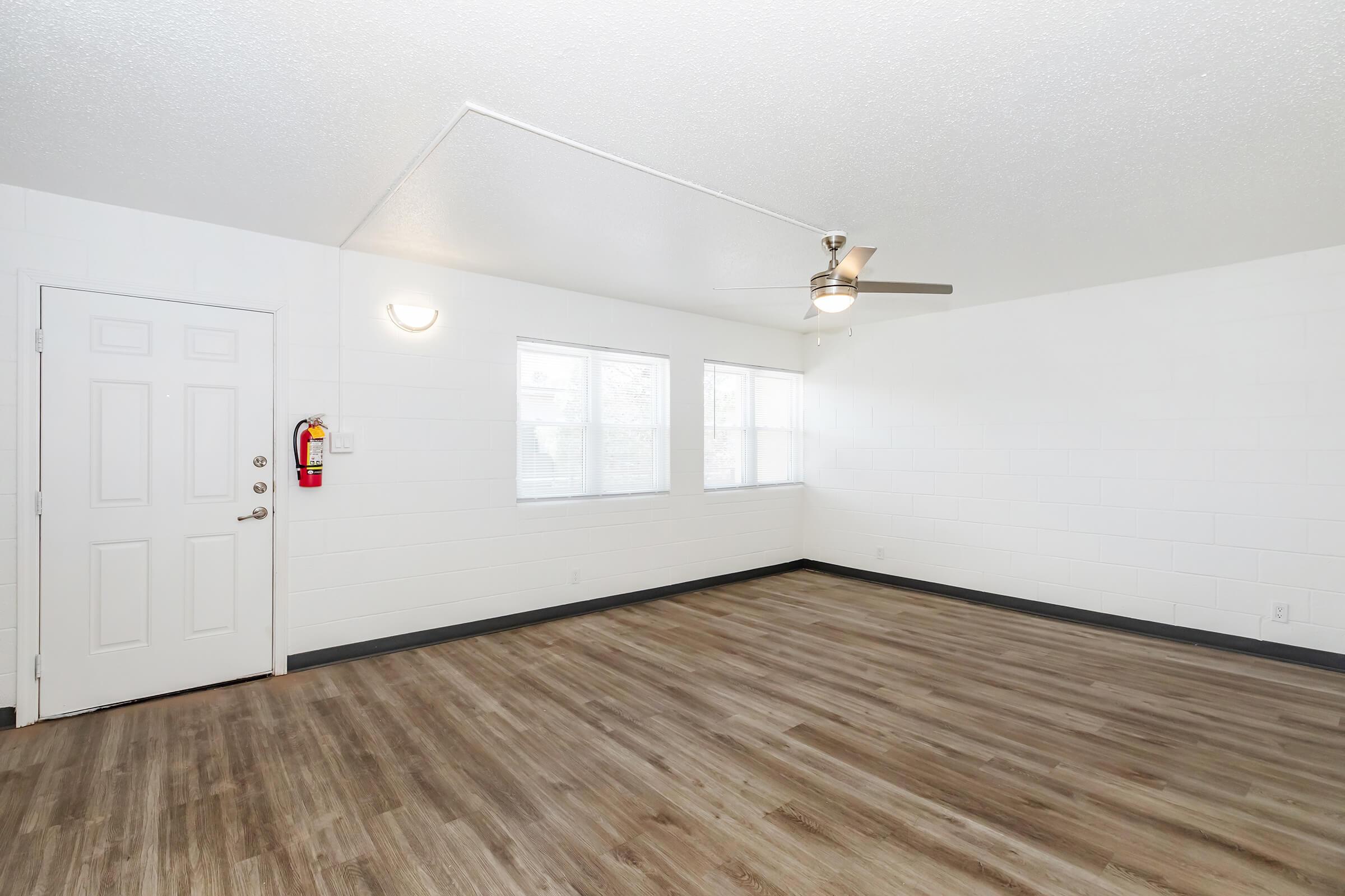 A spacious, empty room featuring light-colored walls, a ceiling fan, and large windows allowing natural light. The floor is covered with faux wood planks. A door leads outside, and a fire extinguisher is mounted on the wall near the entrance.