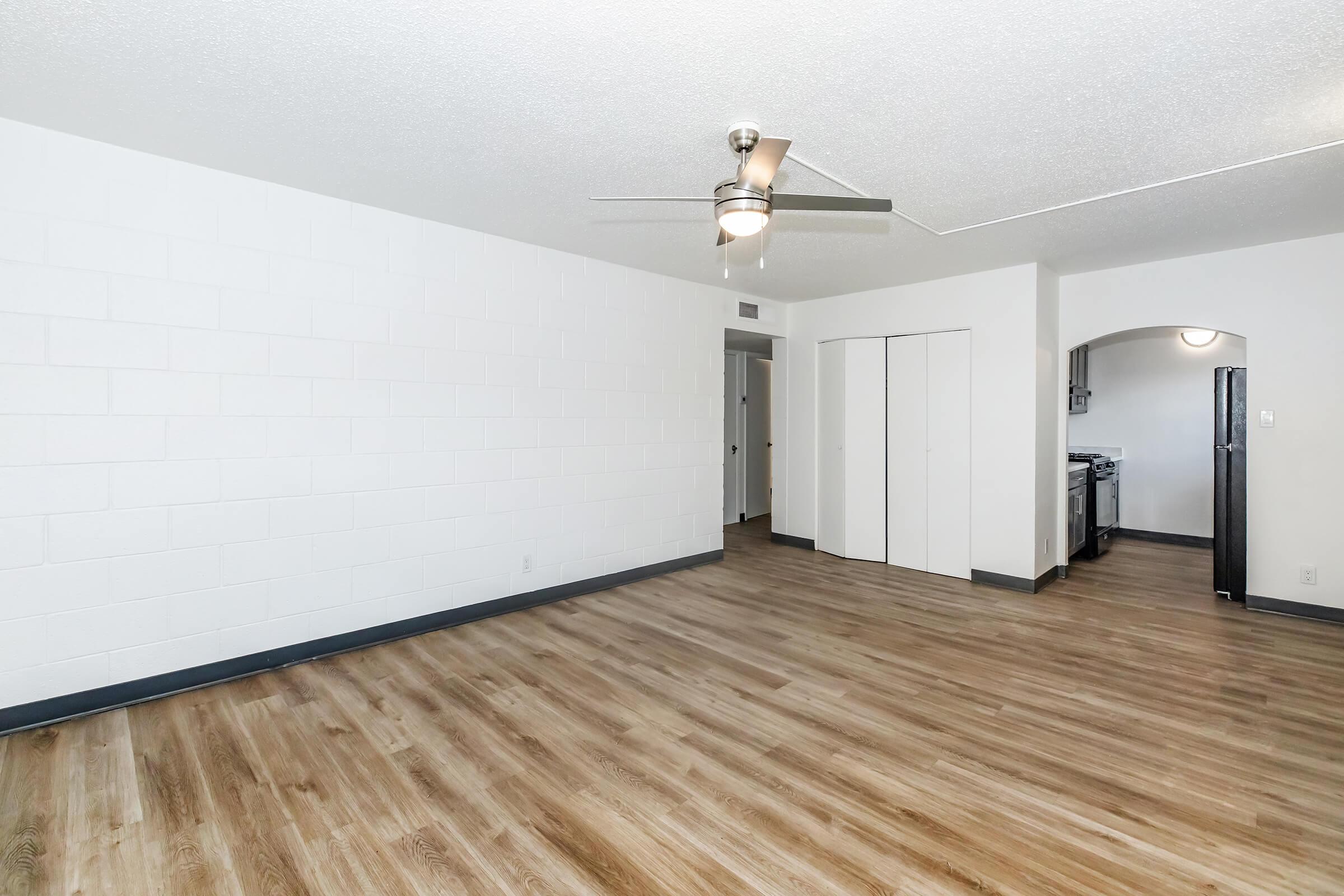 Spacious interior of a modern apartment featuring a light-colored wall, laminated wooden flooring, a ceiling fan, and an open layout. A doorway leads to another room, and a kitchen area is visible in the background with a stove. The space is bright and inviting, ideal for furnishing and personalizing.