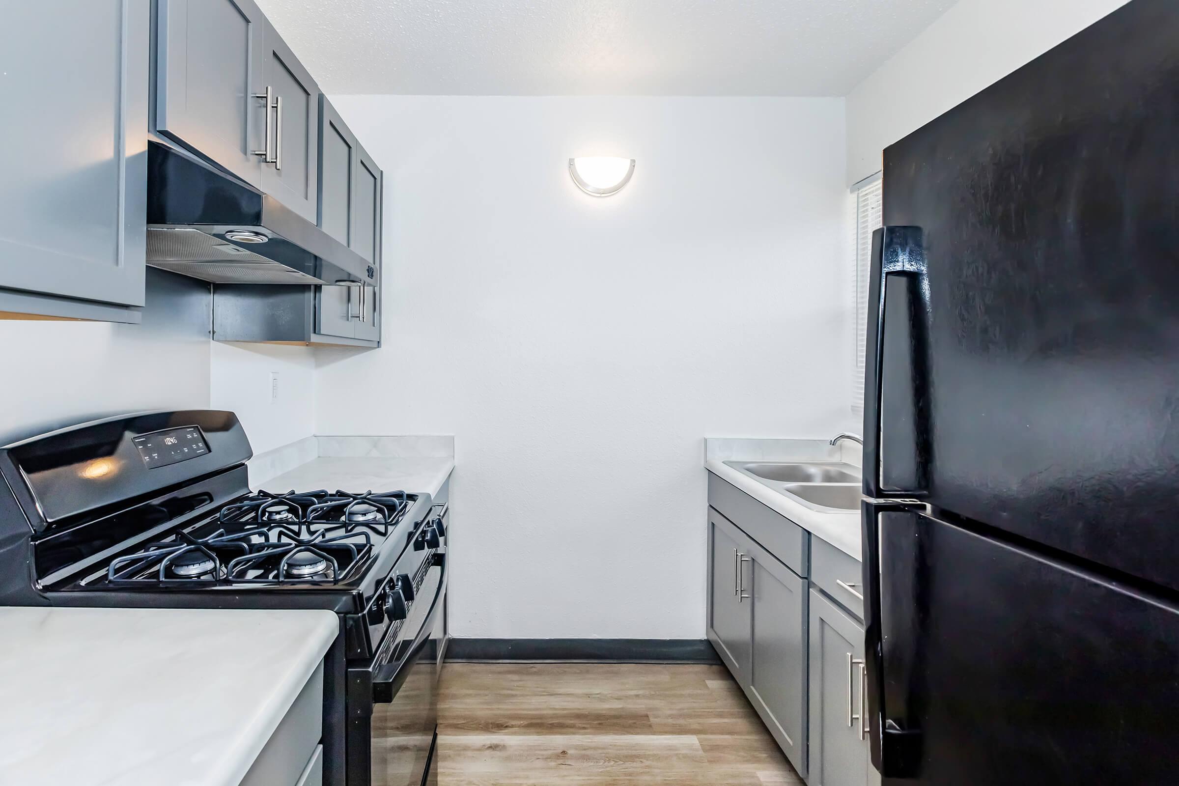 A modern kitchen featuring gray cabinets, a stainless steel gas stove, and a black refrigerator. The countertop is light-colored, and there's a double sink under a window. The walls are painted white, and the space has a warm, inviting atmosphere with wooden flooring.