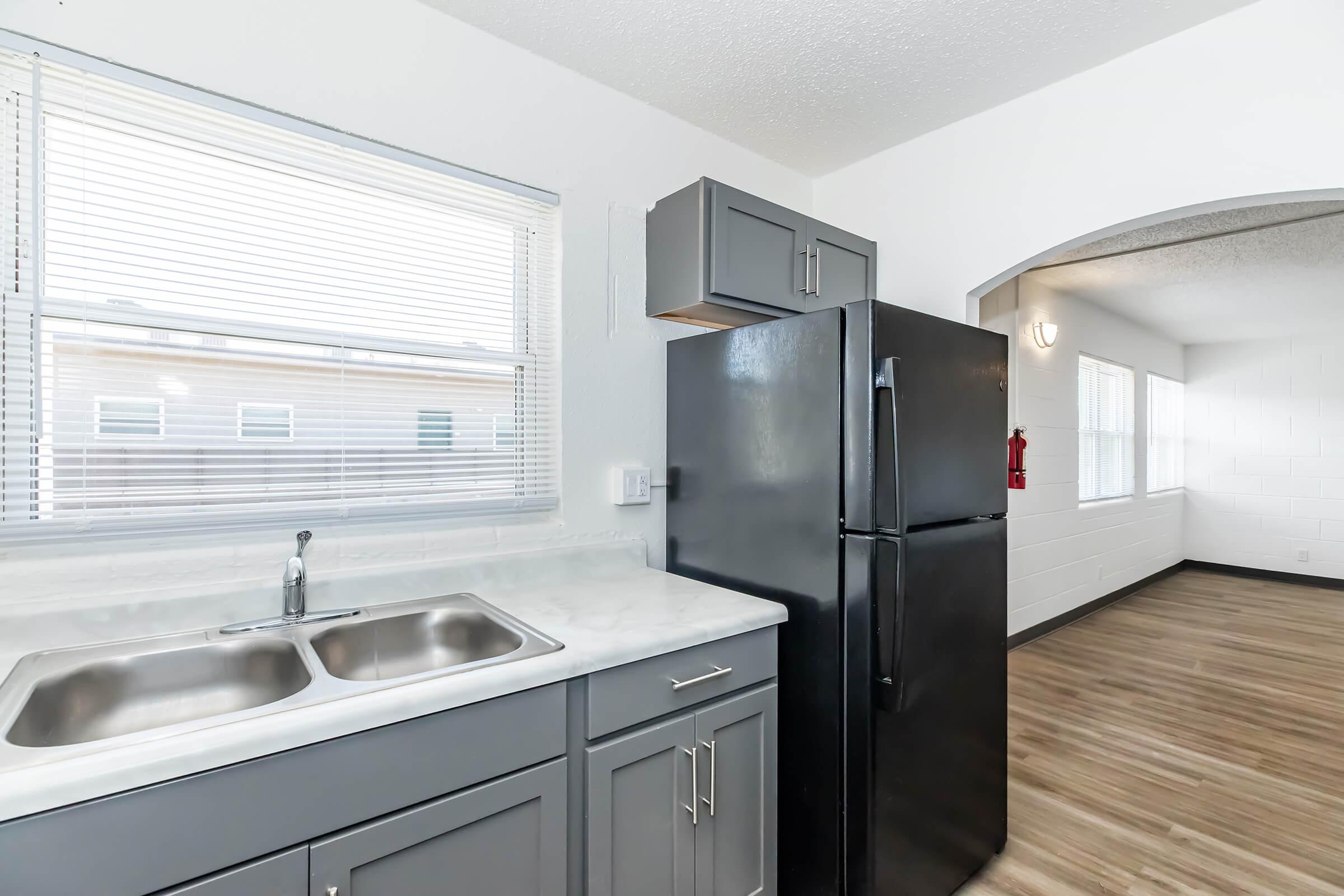 Modern kitchen featuring a dual sink with a marble countertop, gray cabinetry, and a black refrigerator. Natural light filters through a window with blinds. In the background, an archway leads into a brightly lit living space with wooden flooring.