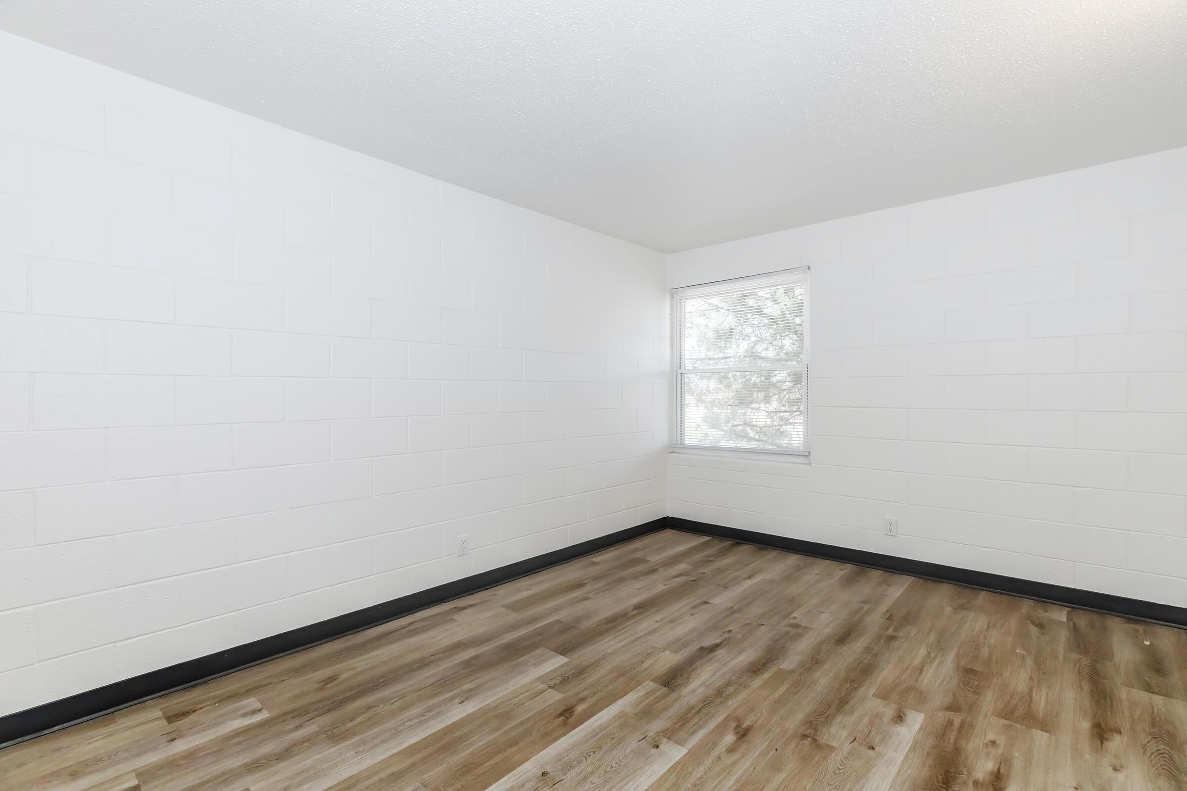 Empty room with white walls and a window allowing natural light. The floor is covered in light-colored laminate. There are no furnishings or decorations, creating a minimalist look. The corner of the room features a slight shadow, and the atmosphere feels bright and airy.