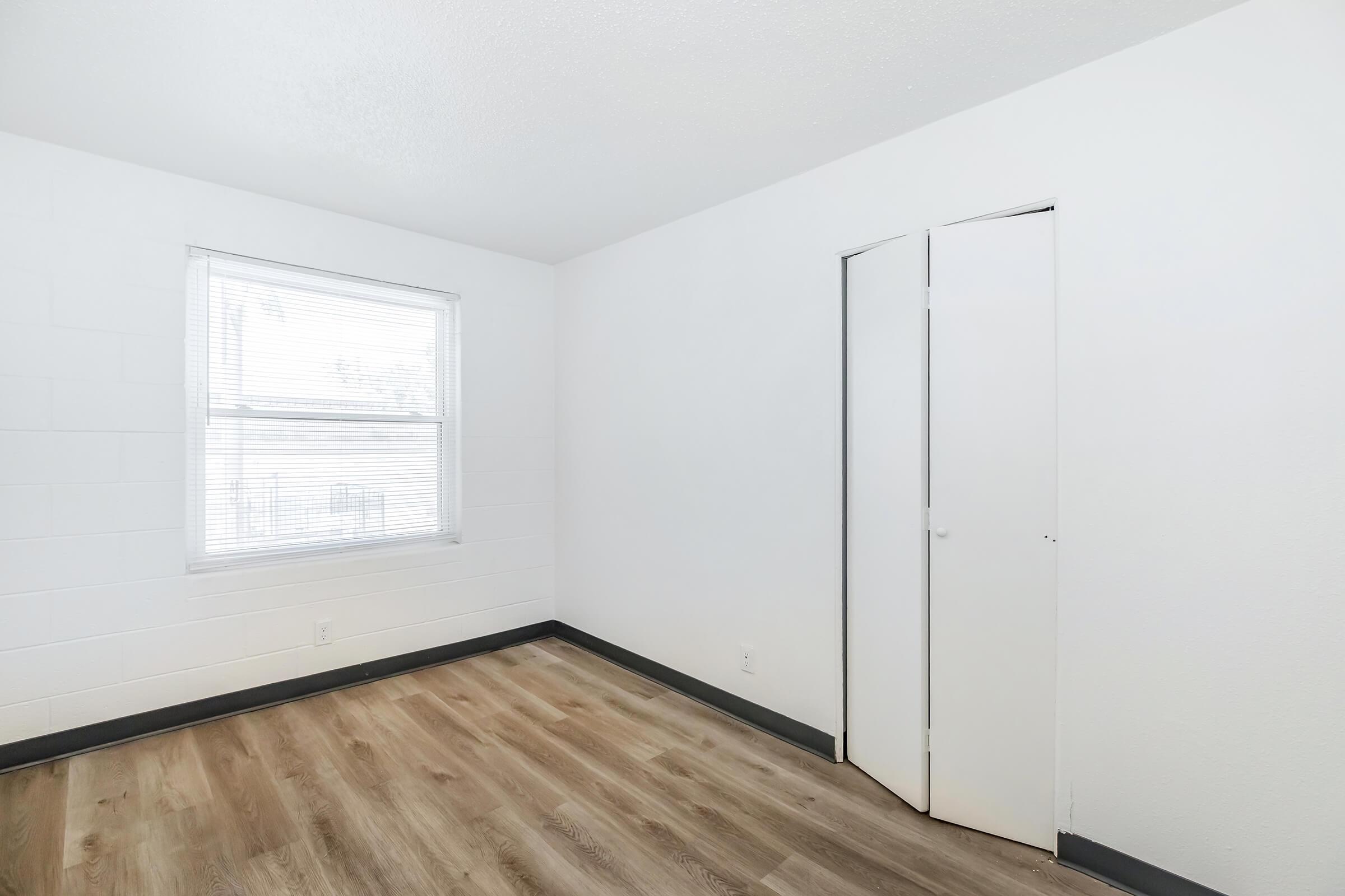 A bright, empty room with light-colored walls and a window featuring blinds. There is a closet with double doors on one side and a wooden floor, enhancing the spacious, minimalist appearance of the space.
