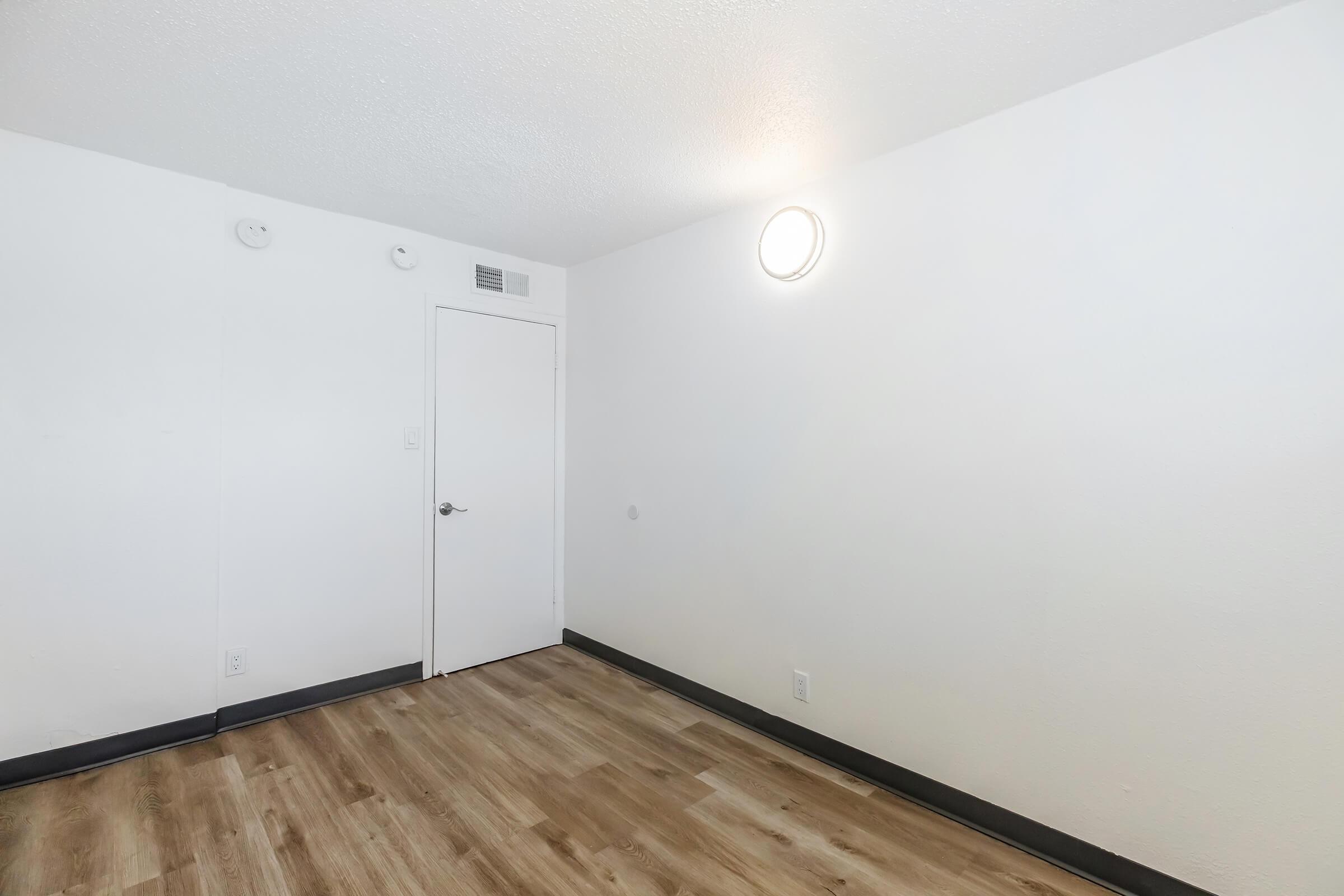 A simple, empty room with white walls, a light fixture on the ceiling, and a closed door. The floor is wooden and has a light brown hue. There are no furnishings or decorations, creating an uncluttered and spacious appearance.
