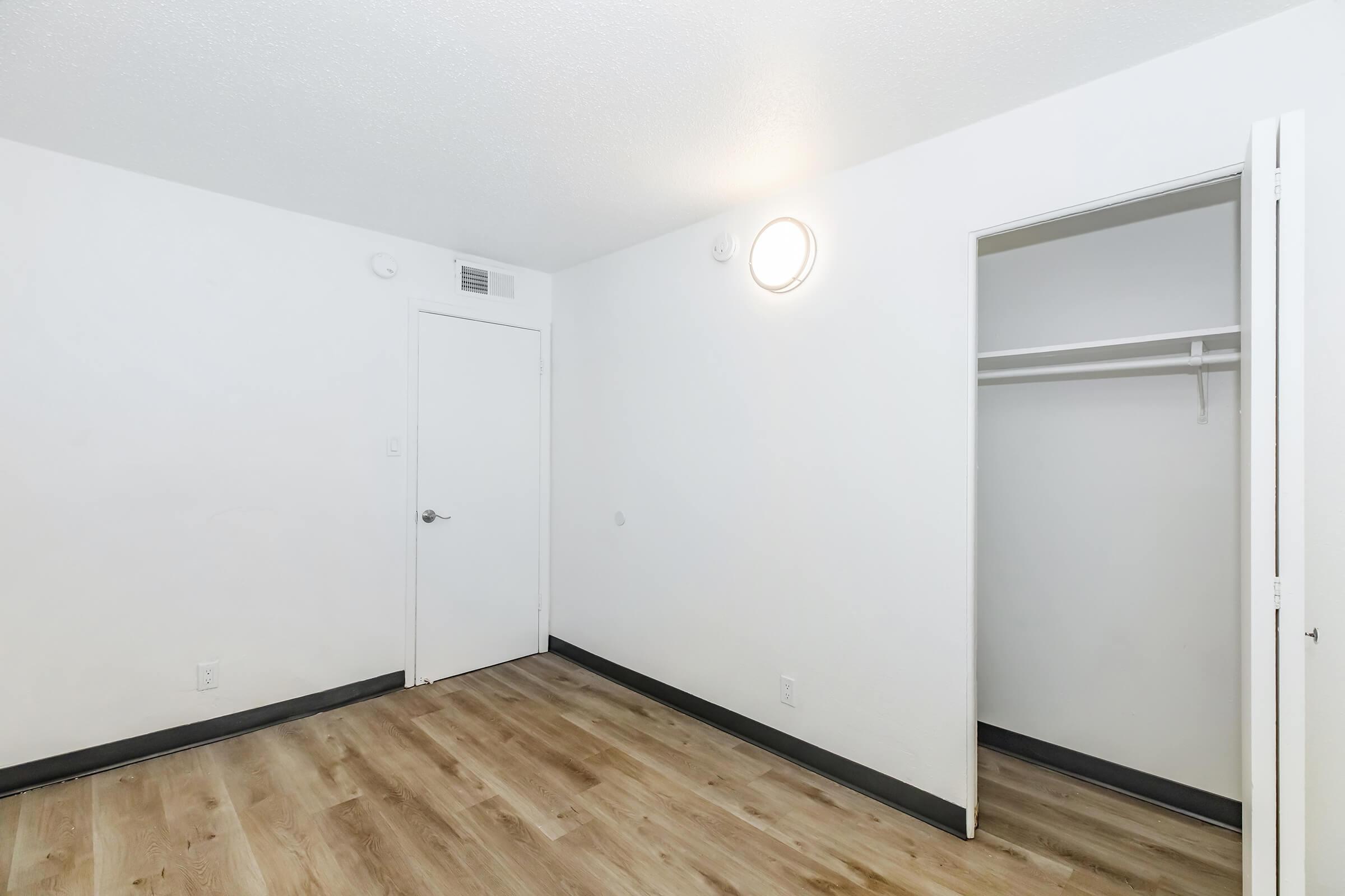 A minimalist room featuring light-colored walls and a wooden floor. There is a door leading to a closet on the right side, a circular ceiling light, and no furnishings. The space has a bright, airy feel, with ample natural light potential.