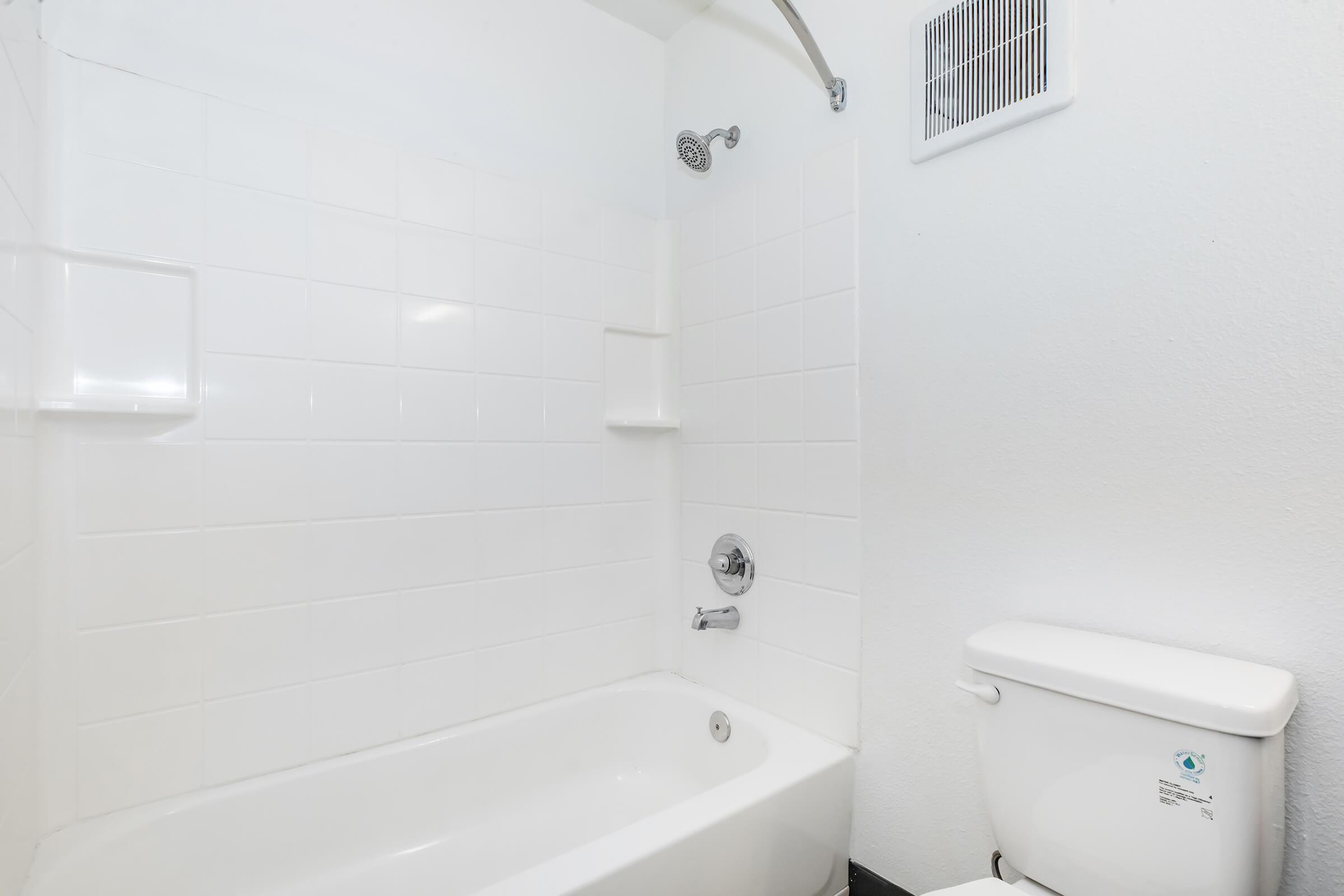 A clean and simple bathroom featuring a white tiled shower area with a bathtub, a wall-mounted showerhead, and a white toilet. The walls are painted white, creating a bright and spacious feel.
