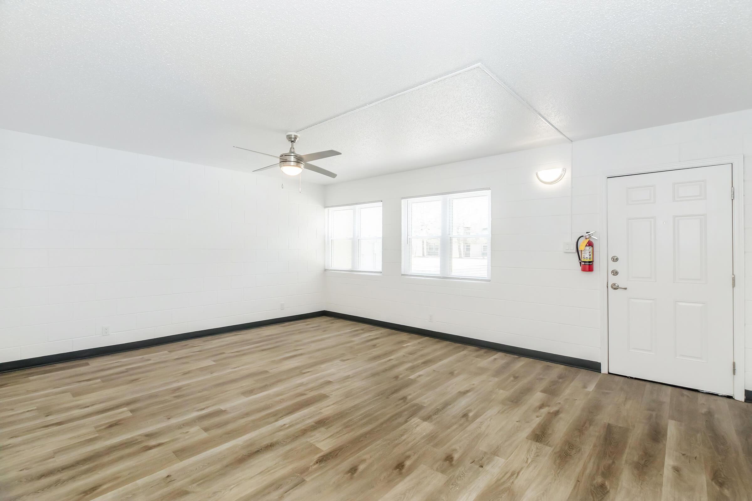 Bright and spacious room with light-colored walls, large windows allowing natural light, and a modern ceiling fan. The floor features warm wood-like laminate, and there is a white door leading outside, complemented by a fire extinguisher mounted on the wall near the entrance.